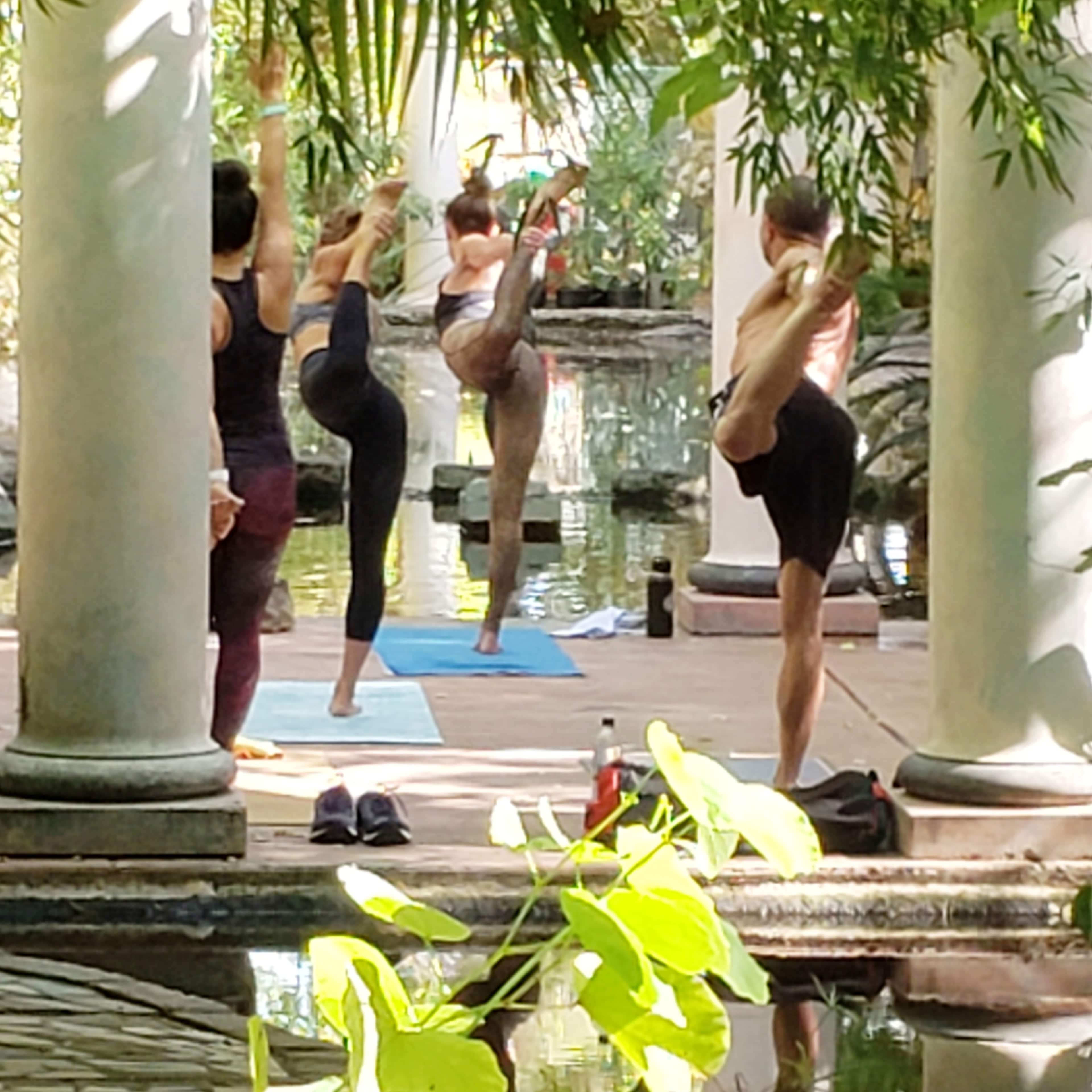 A group of individuals practice yoga near a pond, with columns and greenery surrounding the scene.