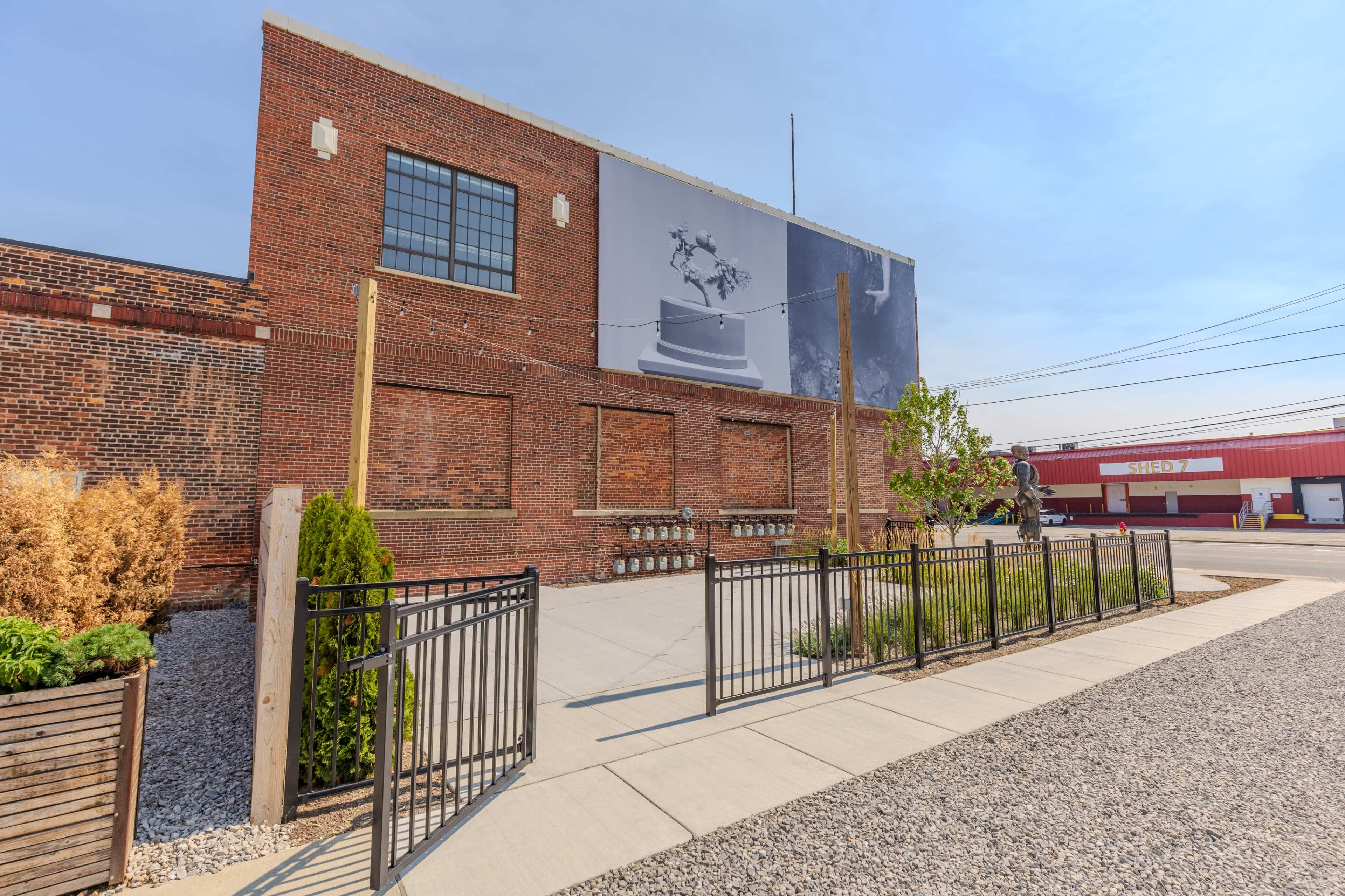 The image shows a brick building with a large mural and a gated landscaped area in front, featuring concrete pathways and greenery.