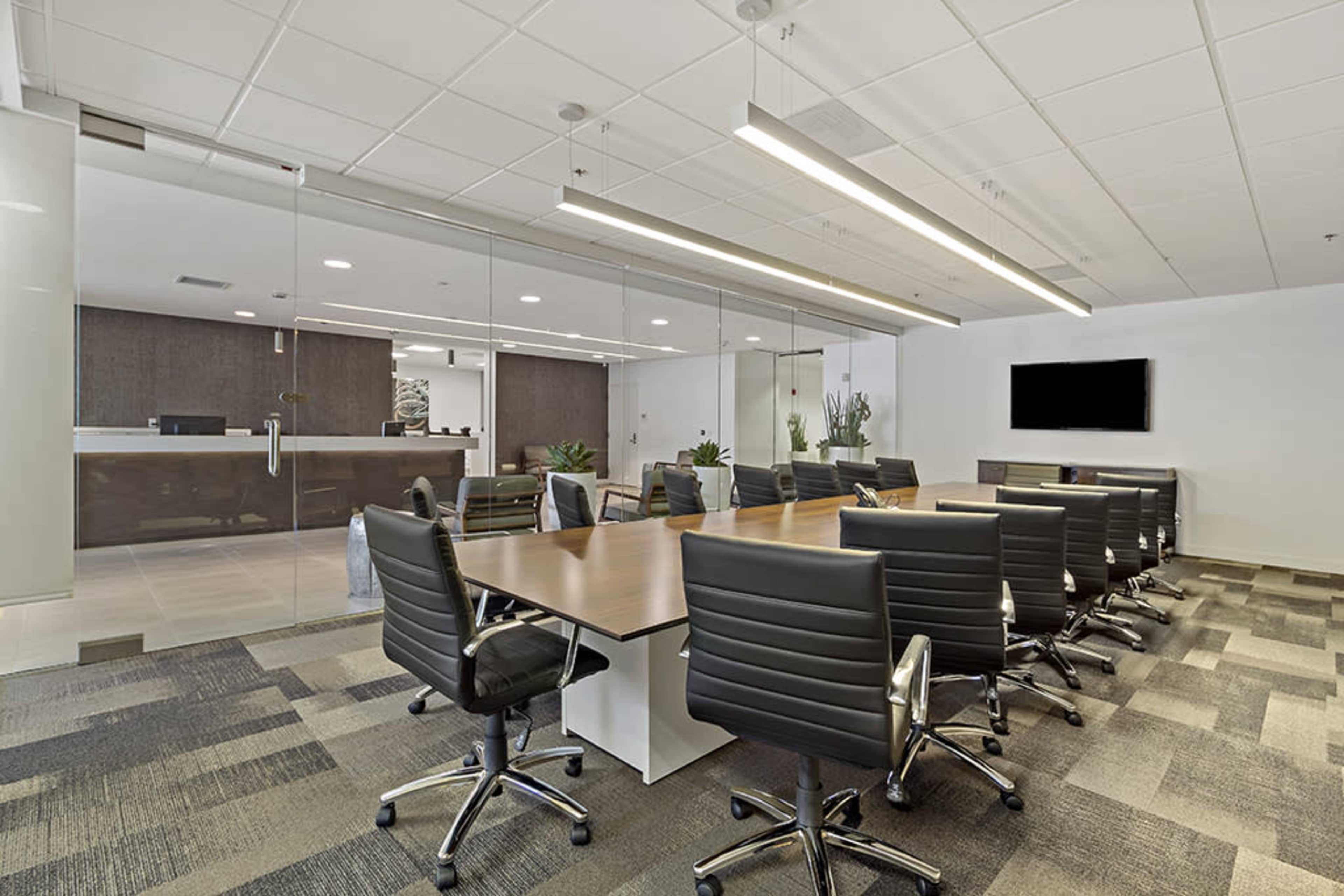 A modern conference room features a long wooden table surrounded by black rolling chairs, with a glass wall overlooking a reception area.