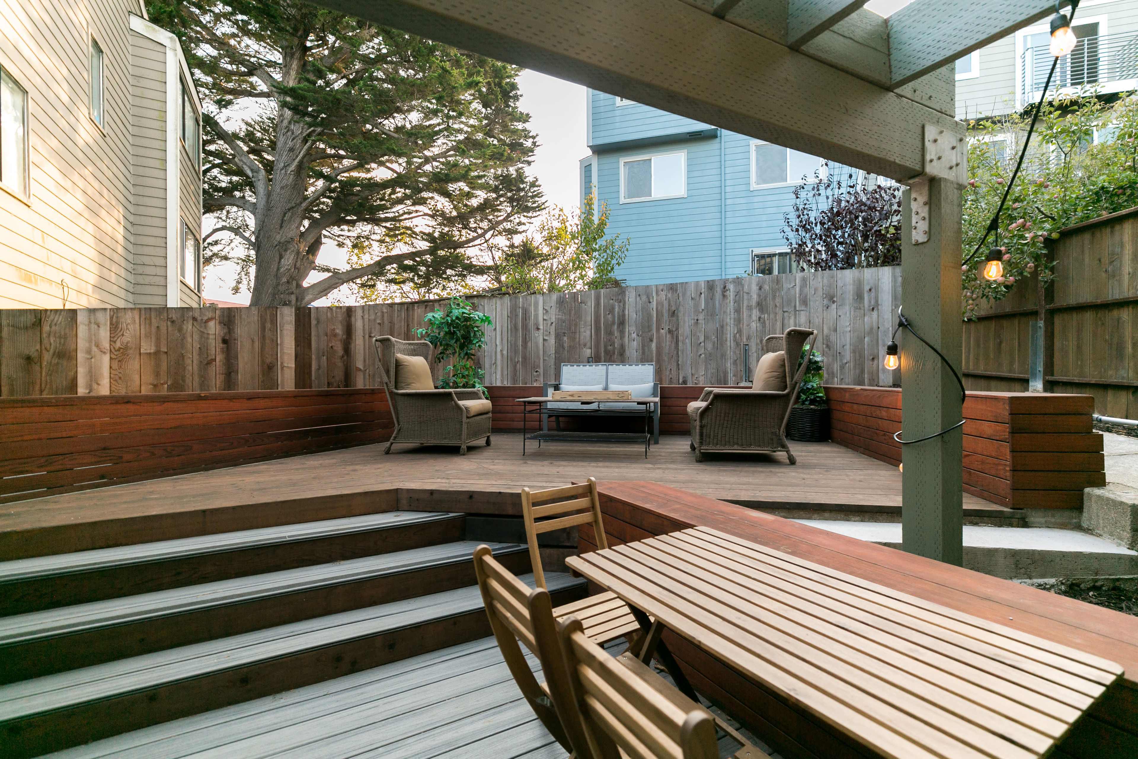 A landscaped backyard with a wooden deck, two armchairs, a coffee table, and a dining table set under a pergola, surrounded by a wooden fence and greenery.