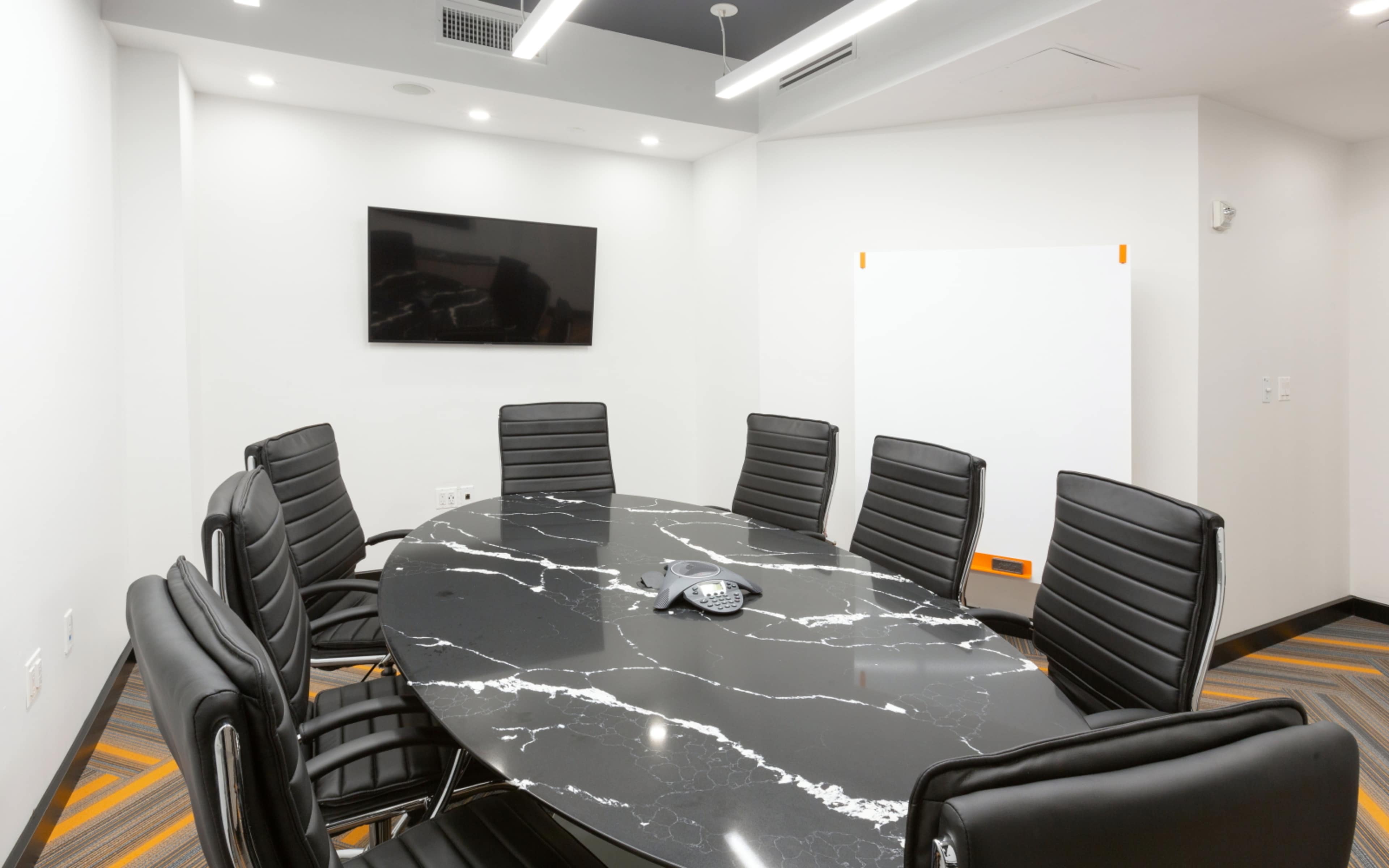 A modern conference room features a large oval black marble table surrounded by eight black leather chairs, with a wall-mounted television and a whiteboard.