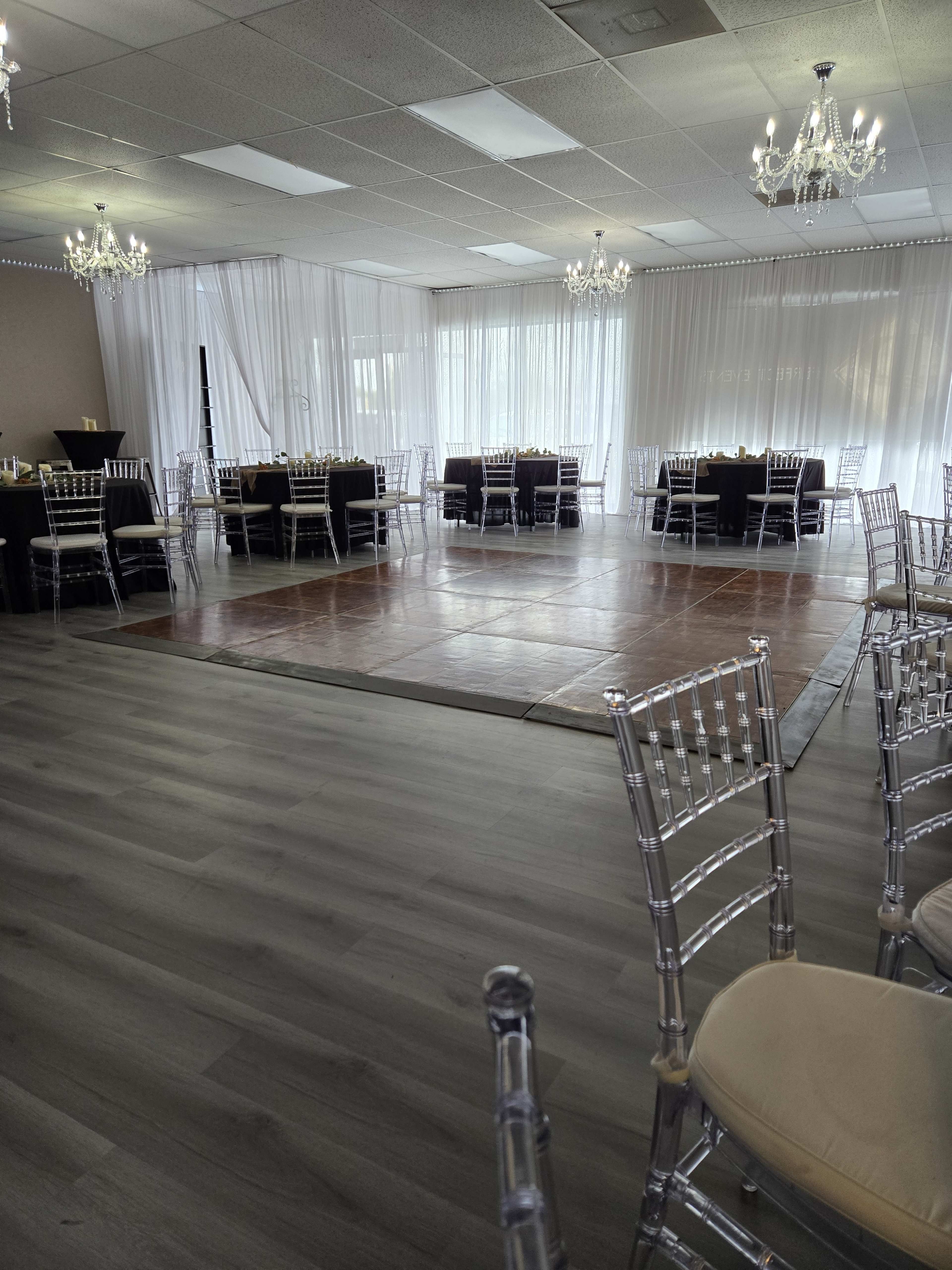 A spacious event venue features arranged tables and chairs with a dance floor, illuminated chandeliers, and sheer drapery.