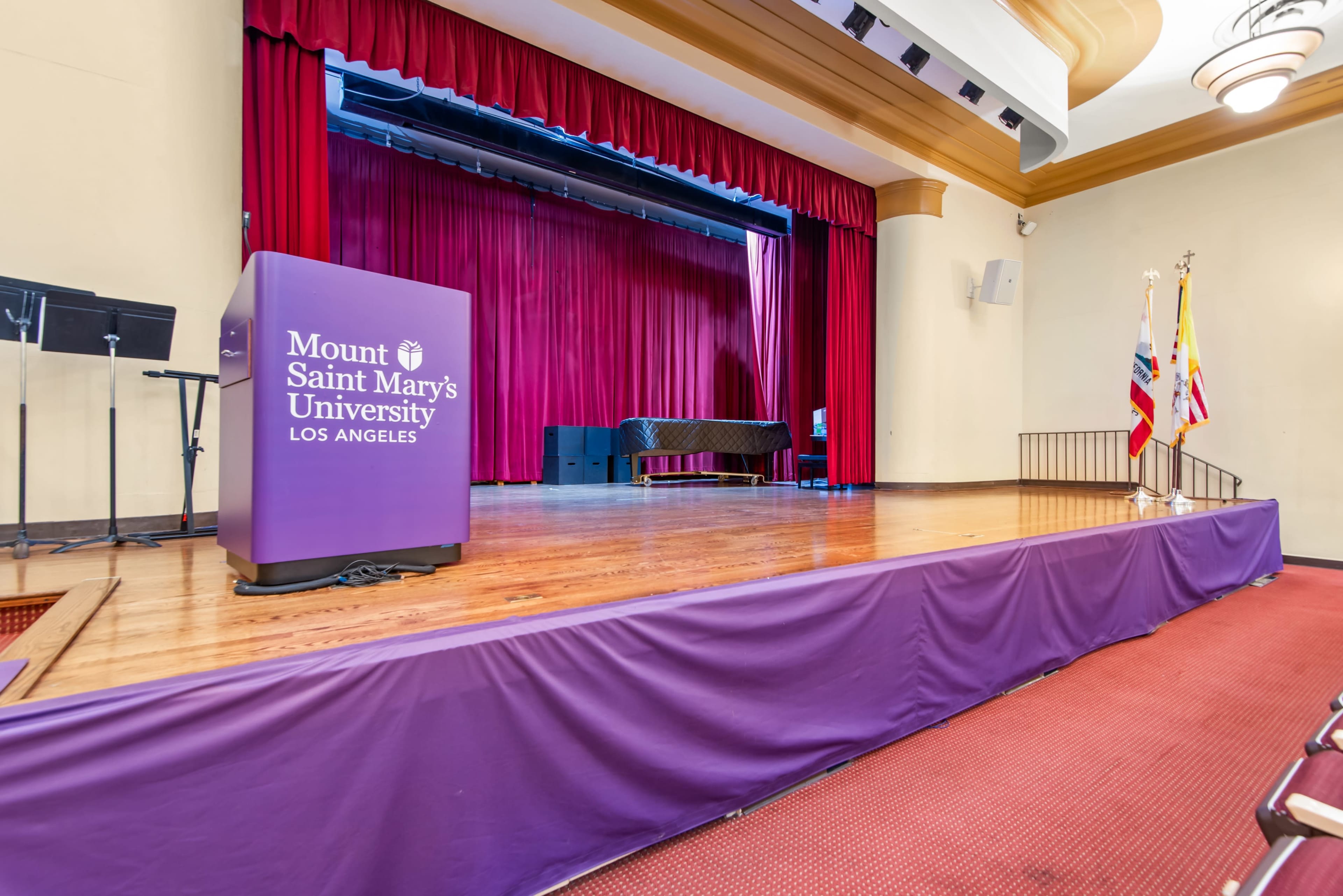 The image shows a stage at Mount Saint Mary's University in Los Angeles, featuring a deep red curtain, a podium, and flags positioned to the side.