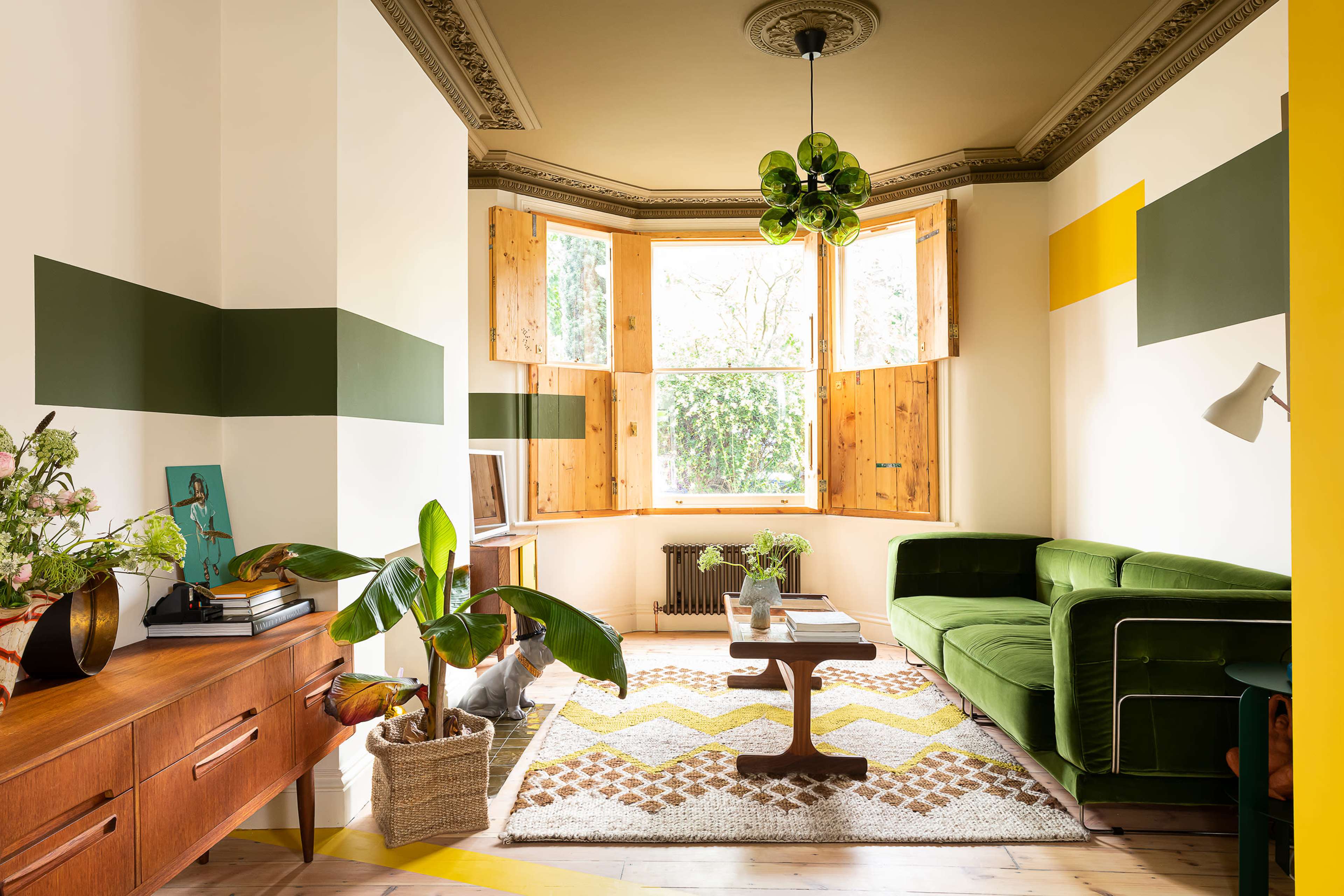A cozy living room features a green sofa, a wooden coffee table, and a window with wooden shutters, surrounded by plants and colorful wall accents.