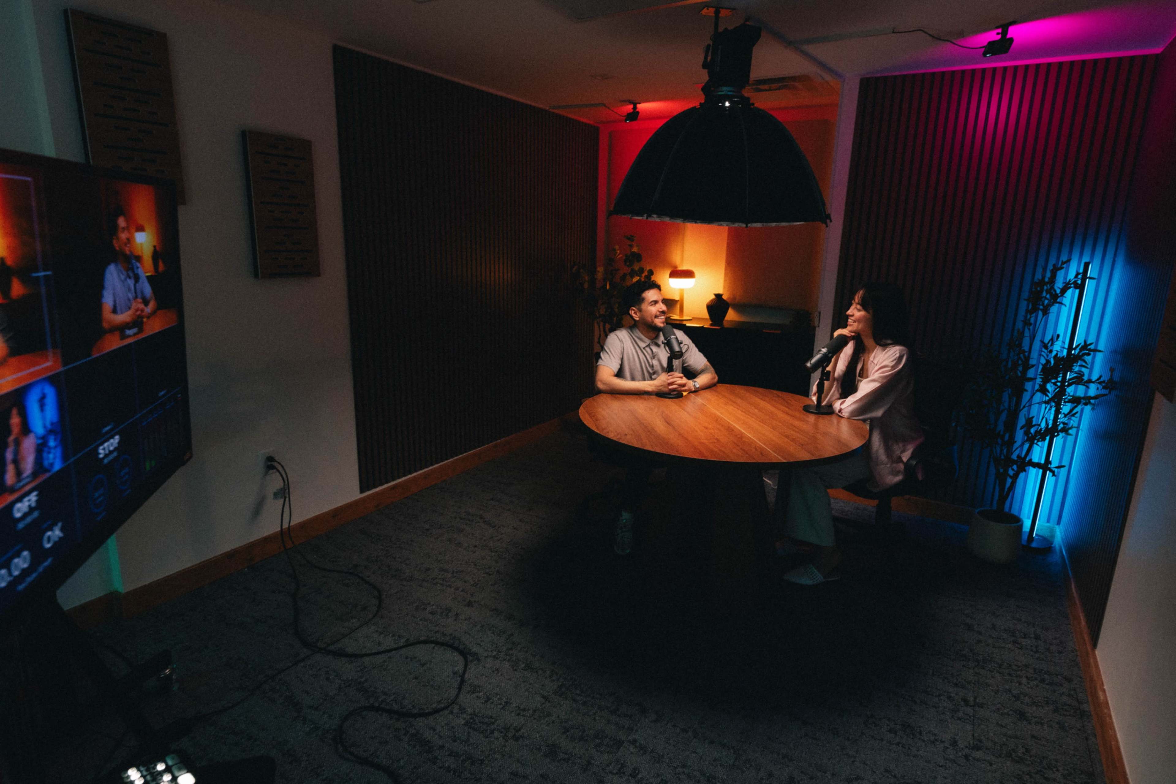 A man and a woman sit at a round table in a well-lit room with colorful lighting and a camera setup nearby.