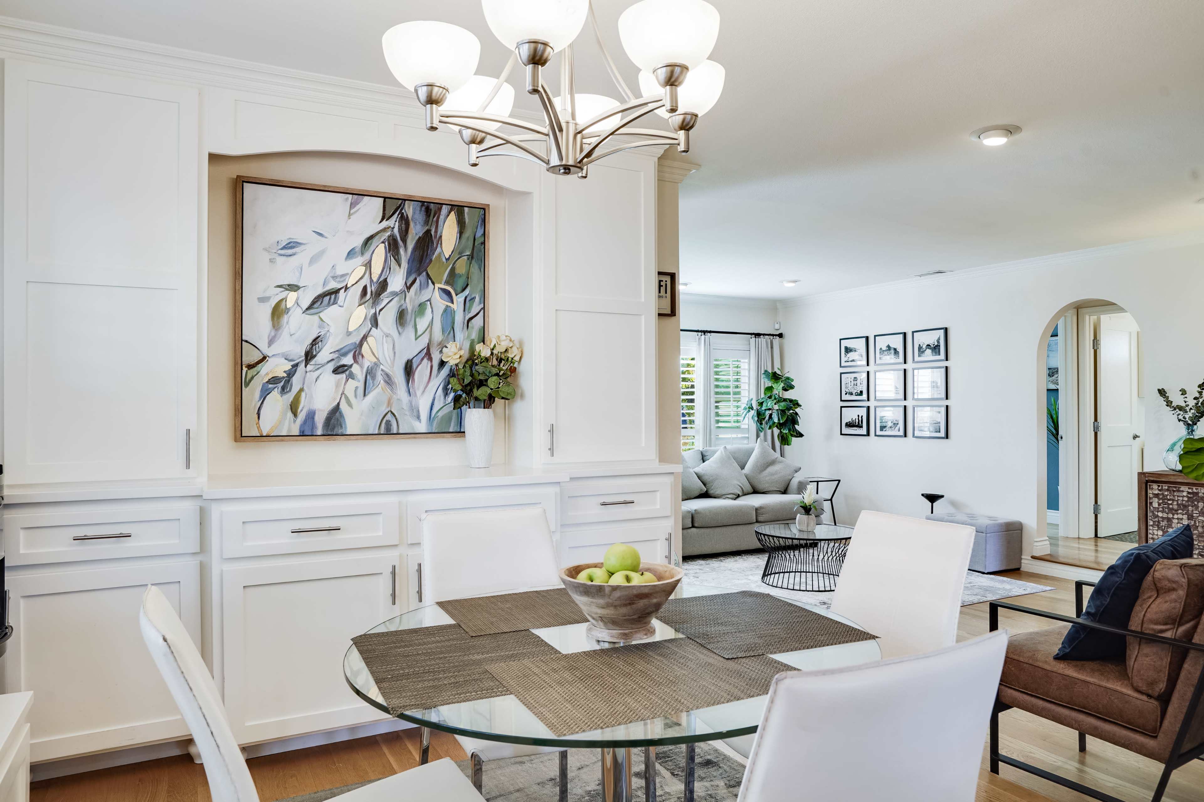 A modern dining area features a round glass table with white chairs, a large artwork on the wall, and open access to a stylish living room.
