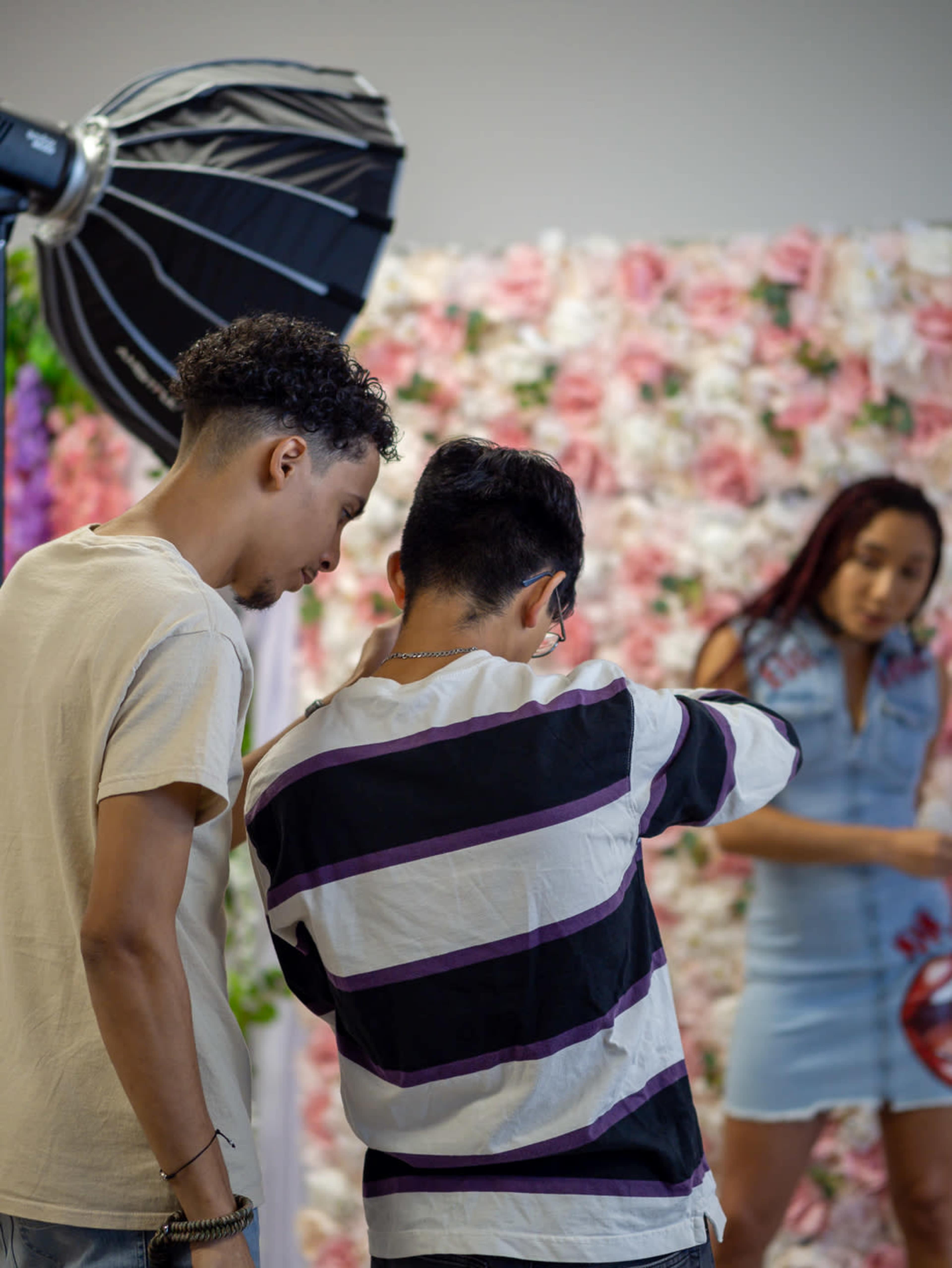 Two young men examine a camera display while a woman in a denim dress poses in front of a floral backdrop.