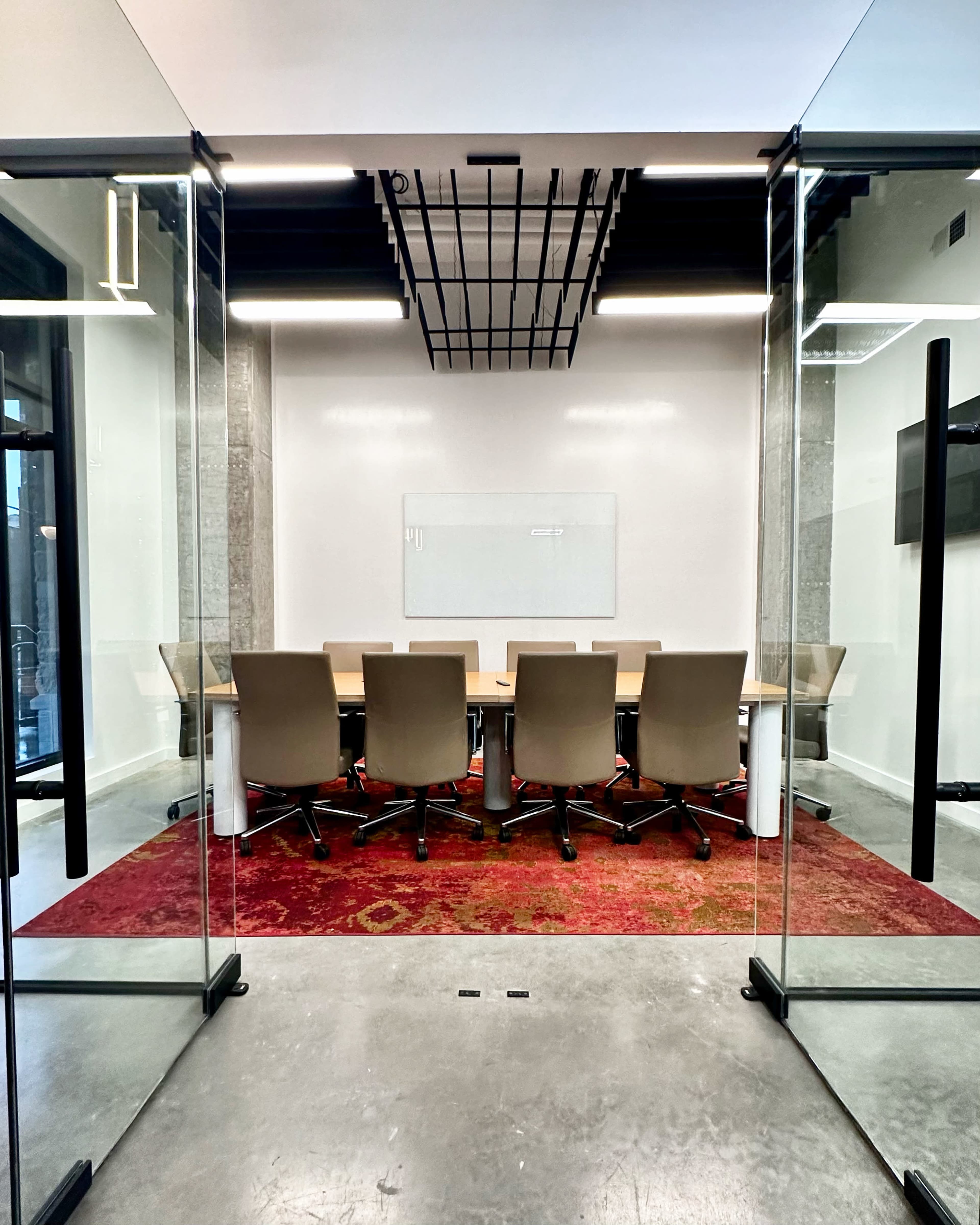 A modern conference room features a long table surrounded by ergonomic chairs on a textured red rug, with large glass walls providing a view into the space.