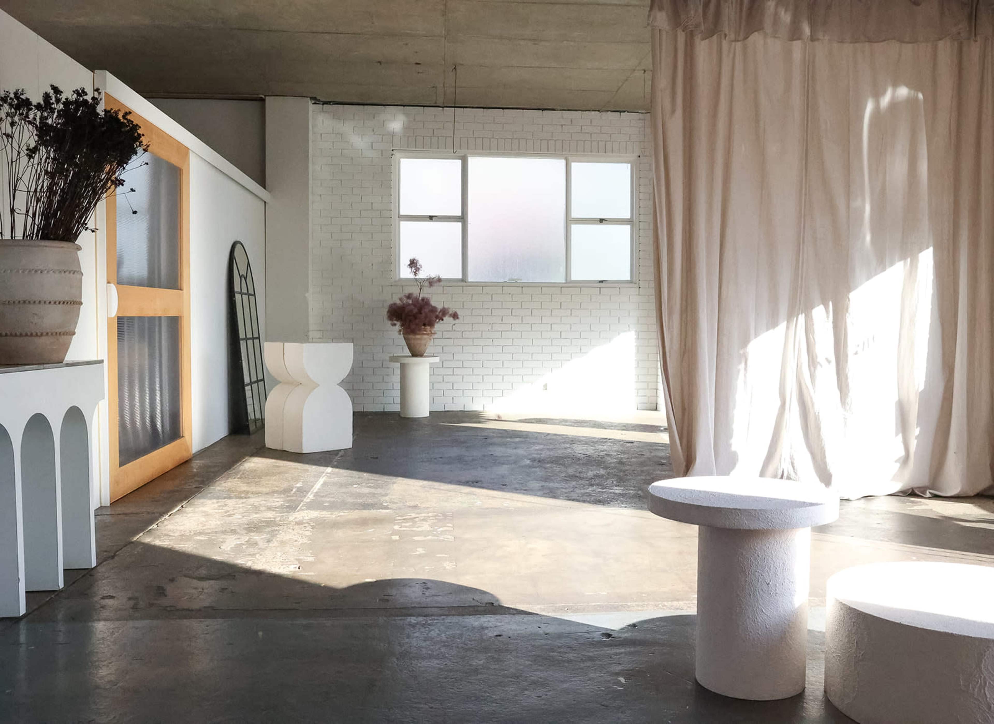 A minimalist interior space with white walls, concrete flooring, and simple decorative elements including a curtain, plants, and geometric furniture.