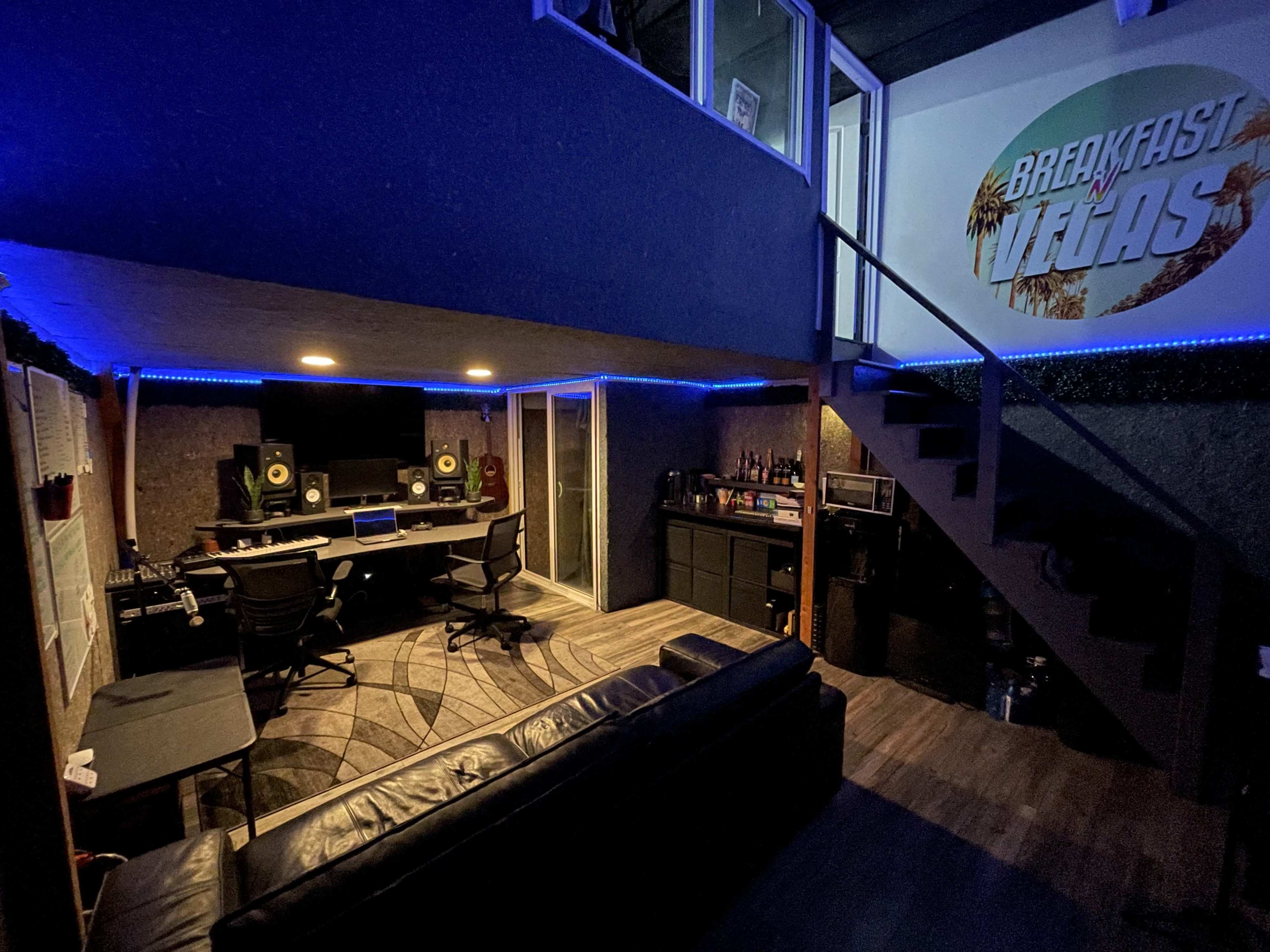 The image shows a dimly lit music studio with a seating area, a recording workstation, and a staircase leading to an upper level.