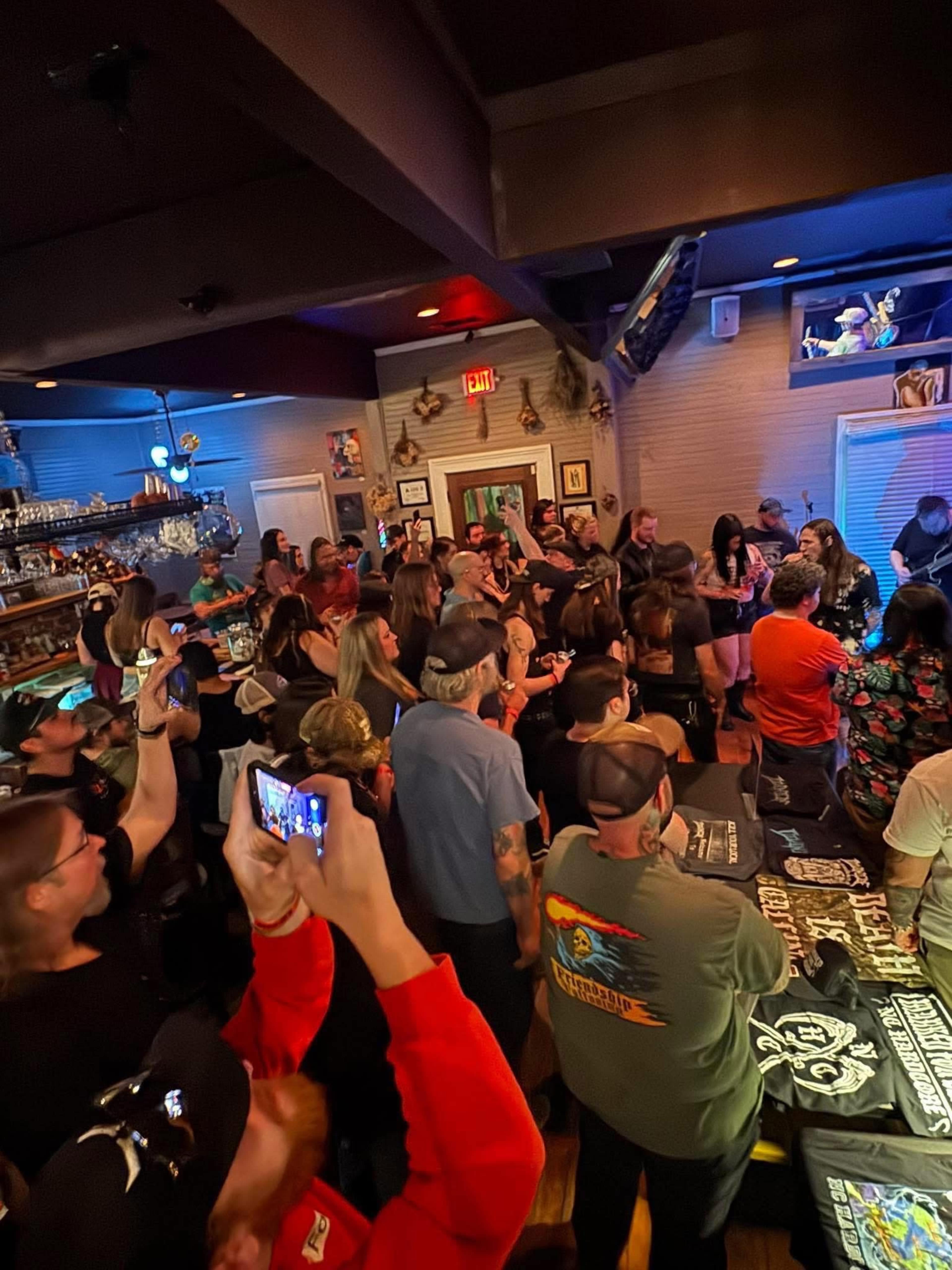 A large crowd is gathered inside a bar, with people standing and socializing around tables while some are recording the scene on their phones.