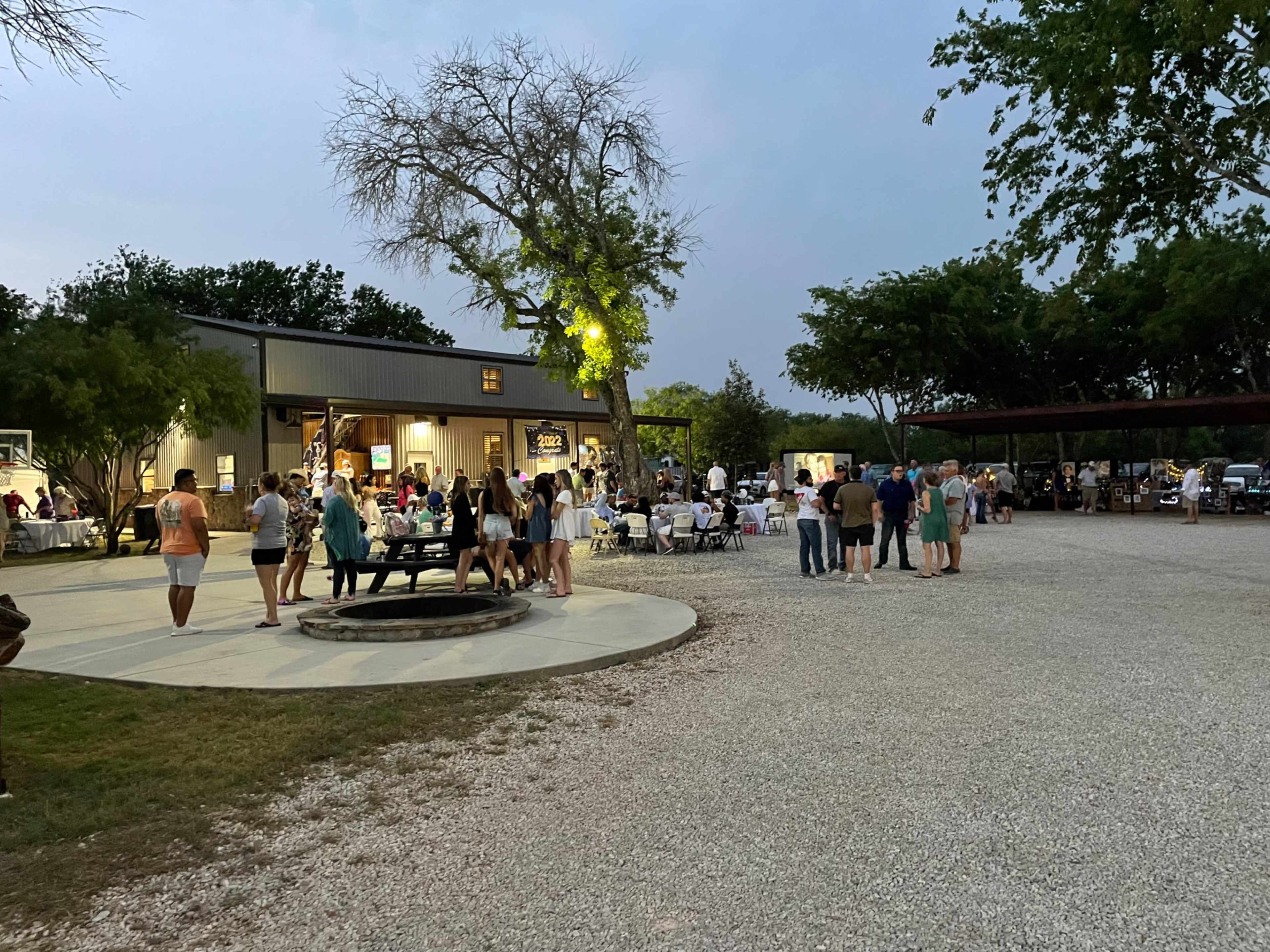 A gathering of people is taking place outside a large building, with tables set up around a fire pit and trees lining the area under an evening sky.
