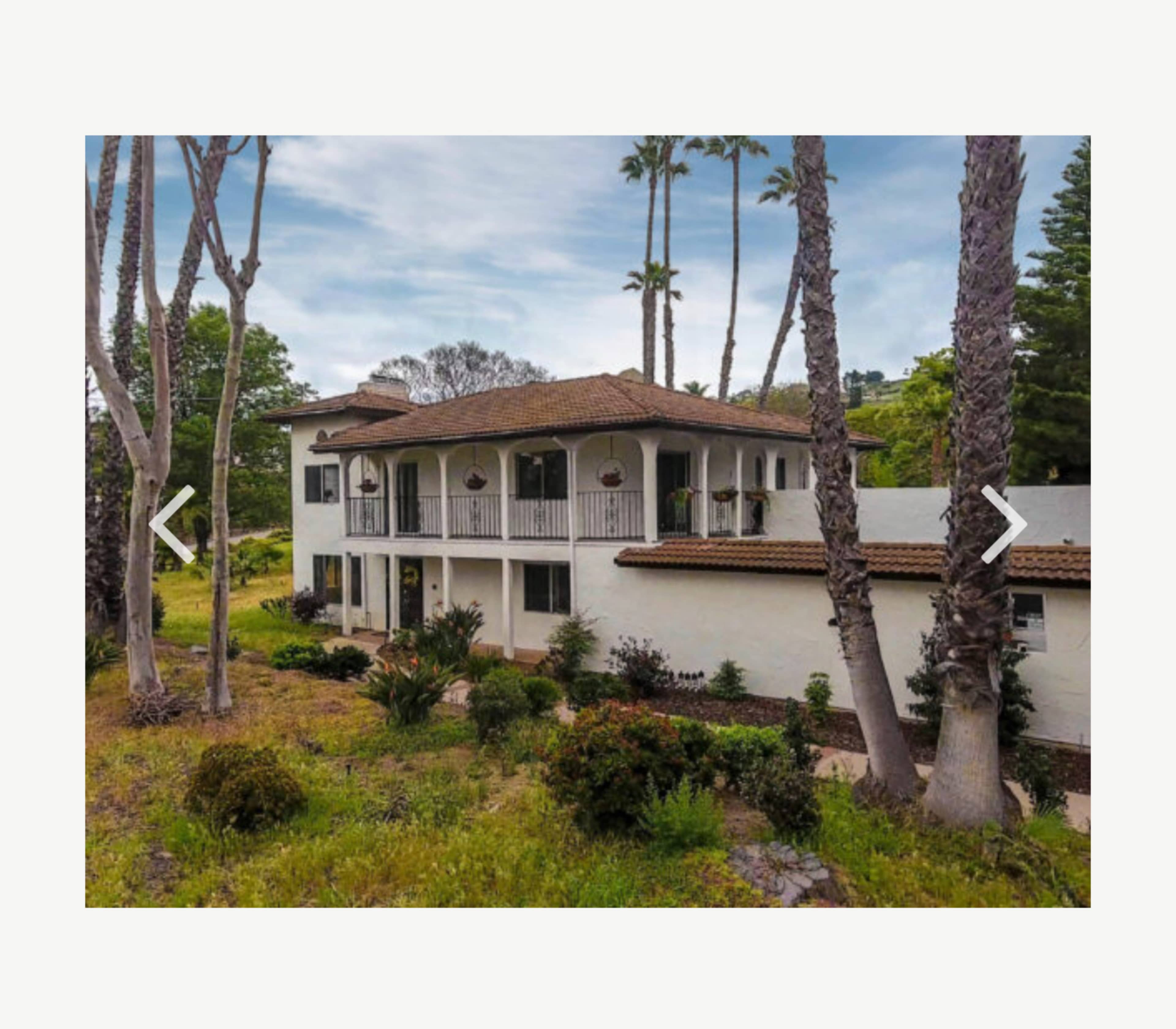 A two-story Mediterranean-style house surrounded by palm trees and lush greenery.