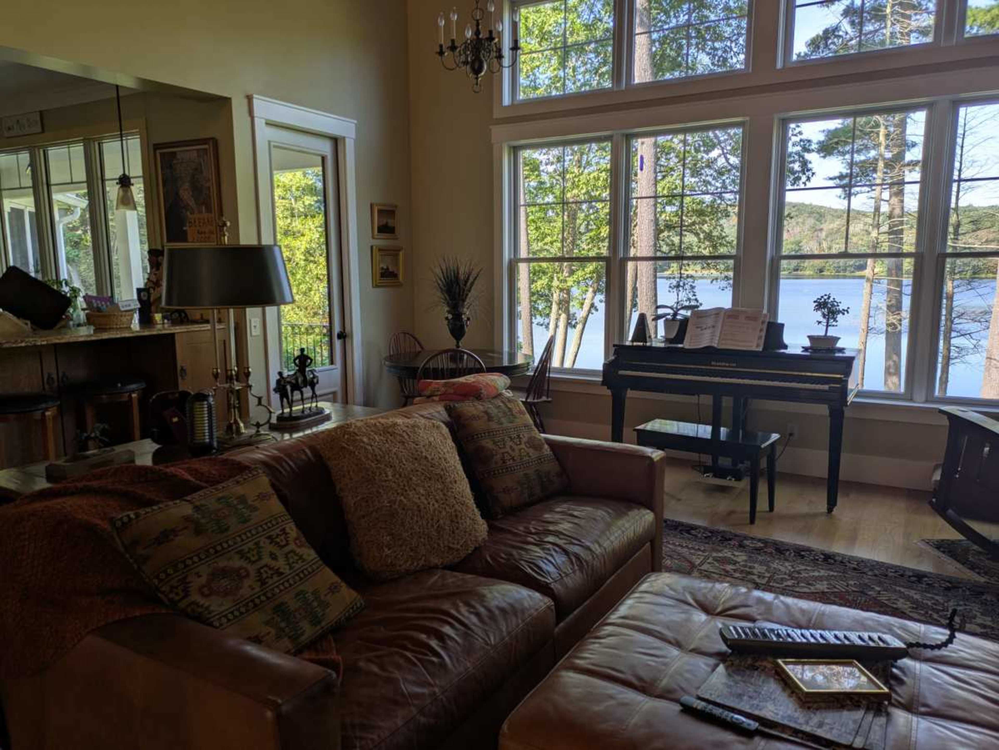 The interior of a cozy living room features a brown leather sofa, a piano near large windows overlooking a lake and trees.