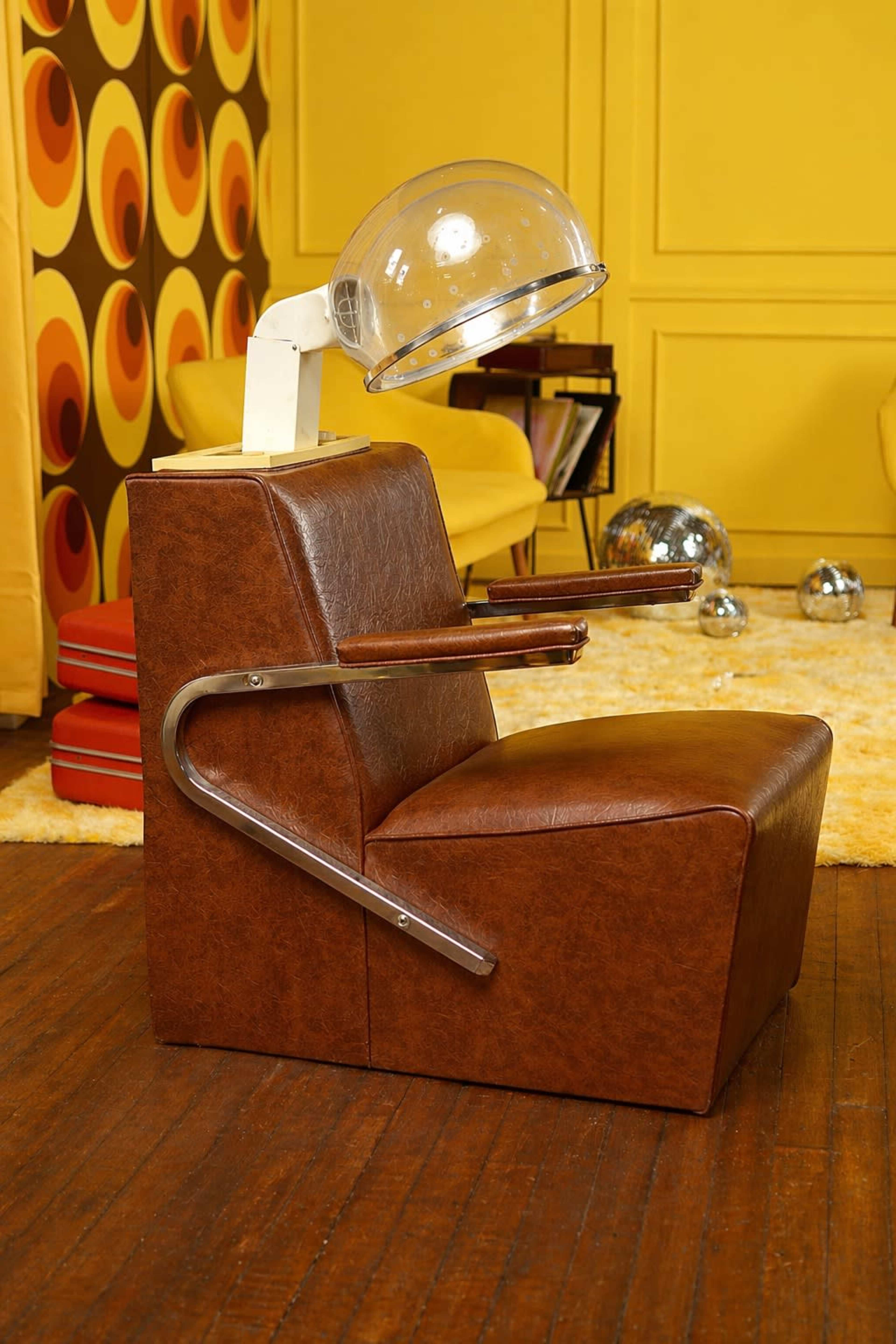 The image shows a vintage salon chair with a clear dome-shaped hair dryer attached, set against a backdrop of retro patterned wallpaper and brightly colored furnishings.