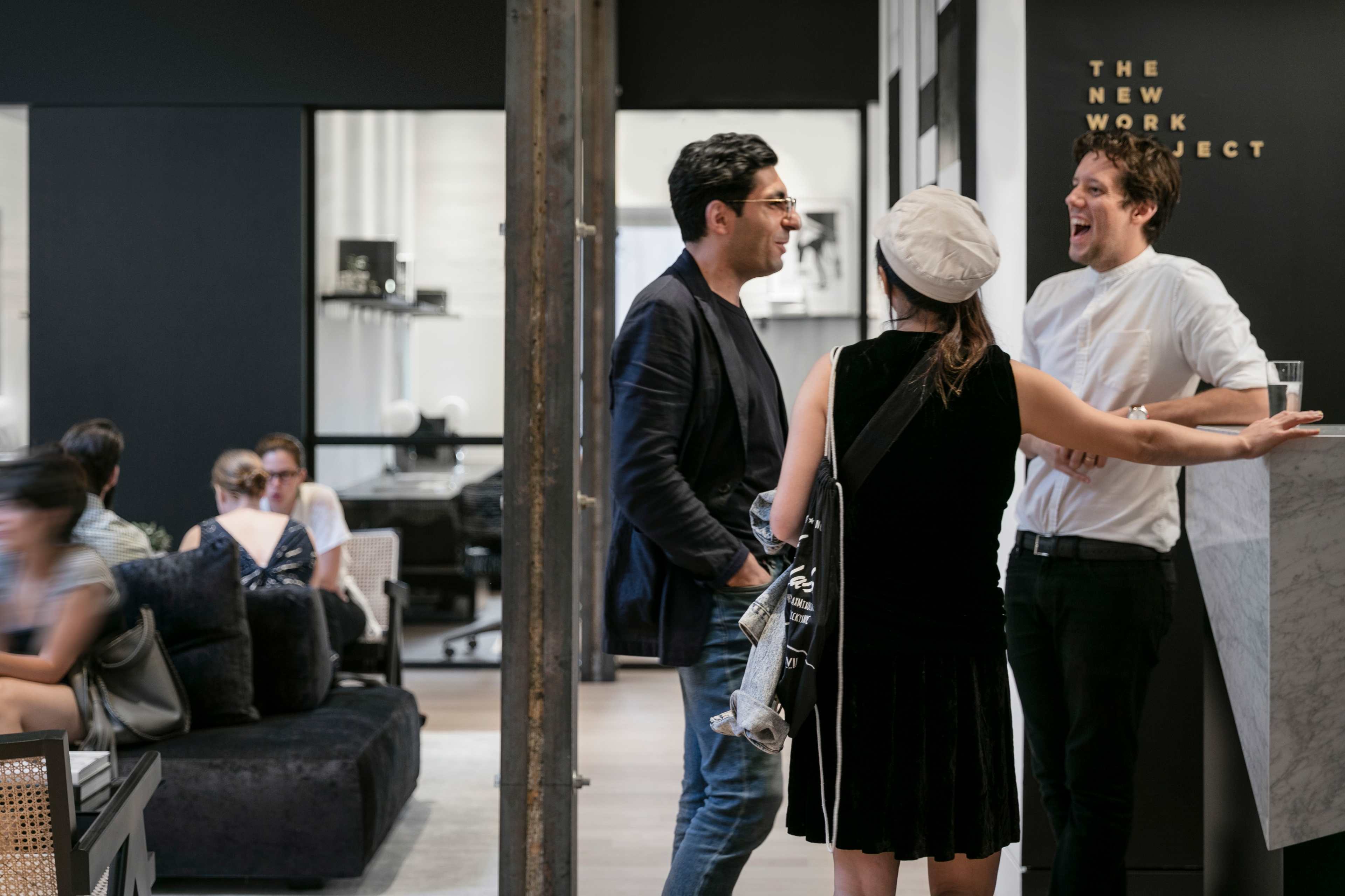 Three people engage in conversation in a modern office space, while others are seated in the background.