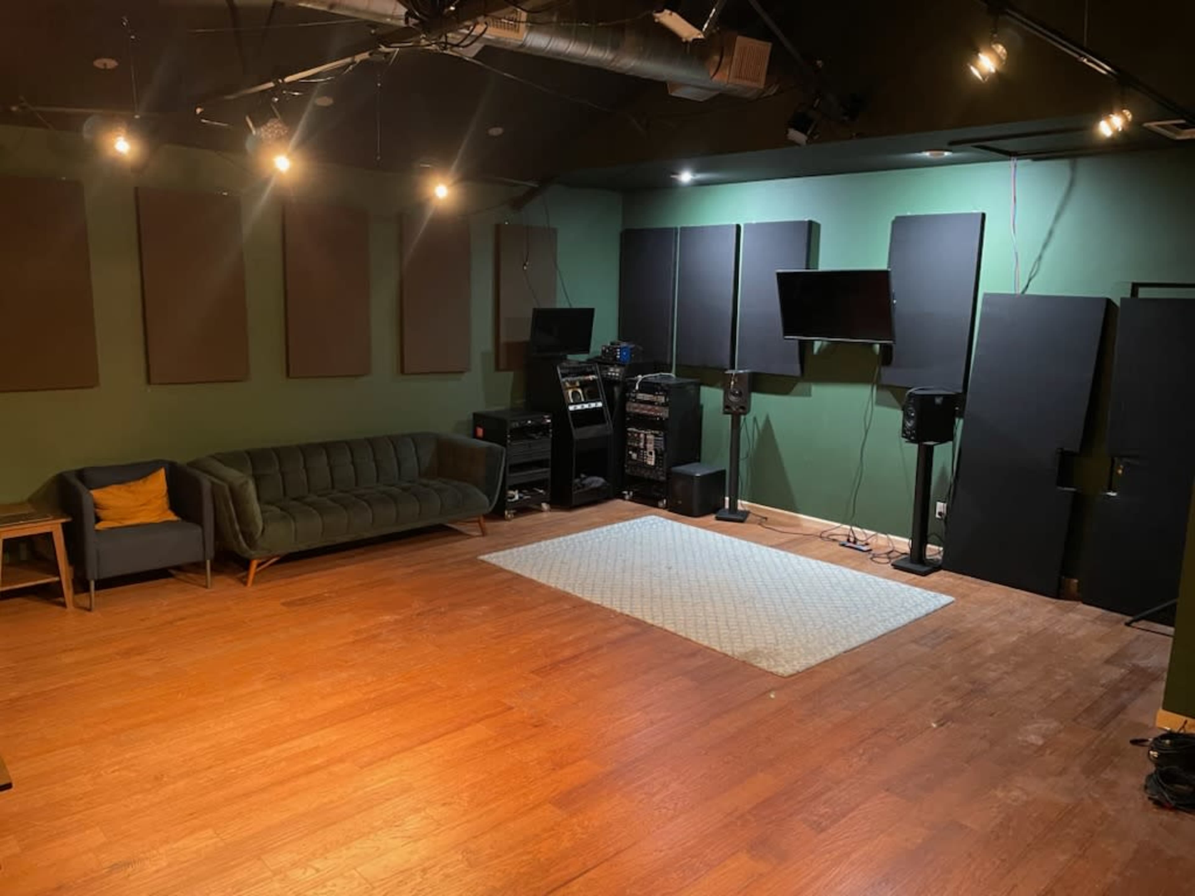 A sound studio with a green wall, wooden floor, a dark sofa, audio equipment, and soundproof panels.