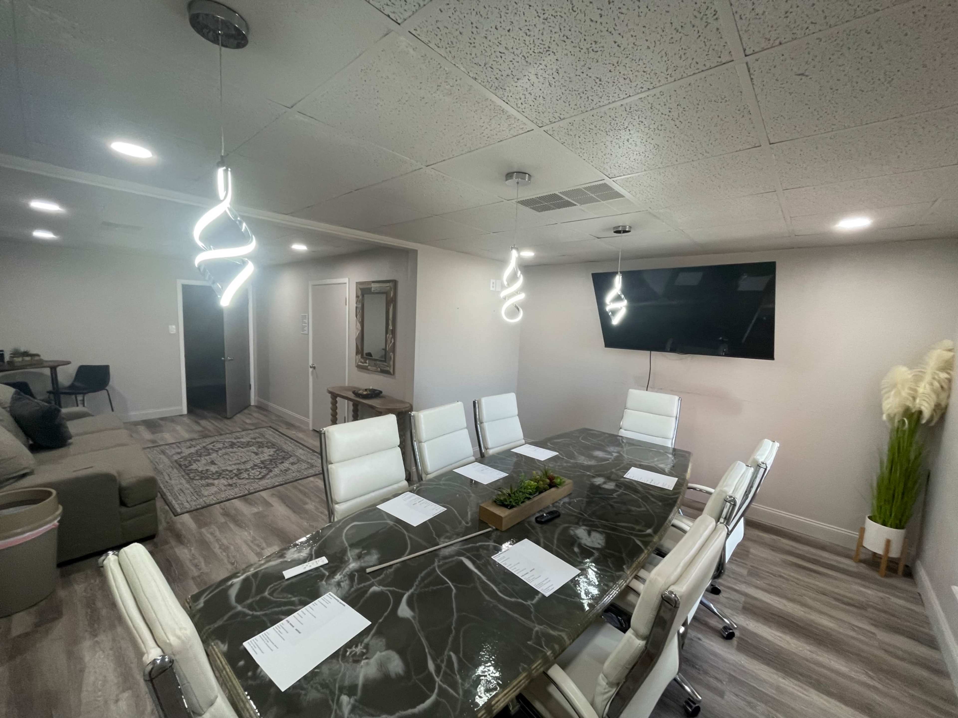 A modern conference room features a long marble table surrounded by white chairs, with a large screen mounted on the wall and decorative lighting overhead.