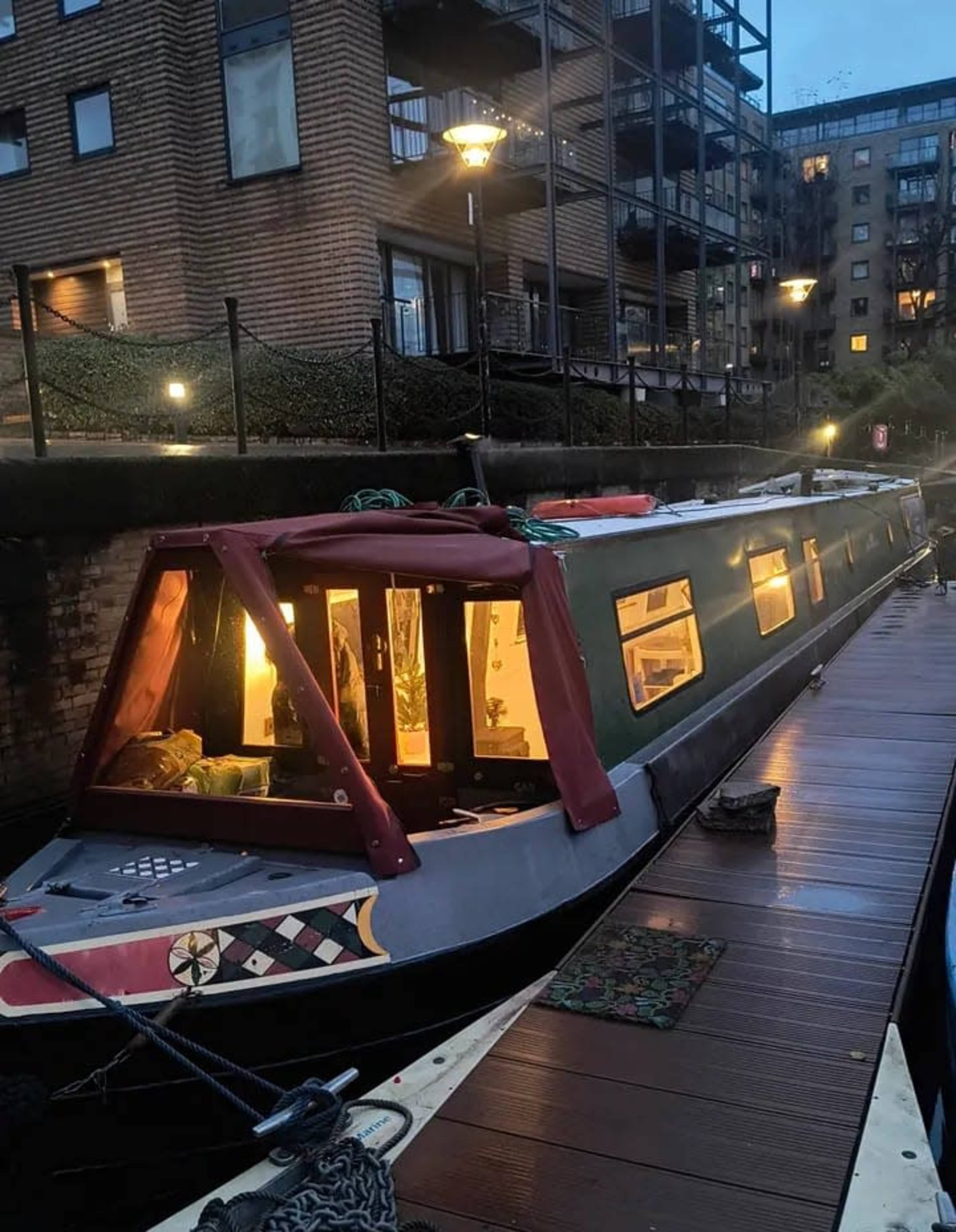 A narrowboat is moored beside a wooden dock, illuminated from within, while nearby residential buildings and streetlights are visible in the background.