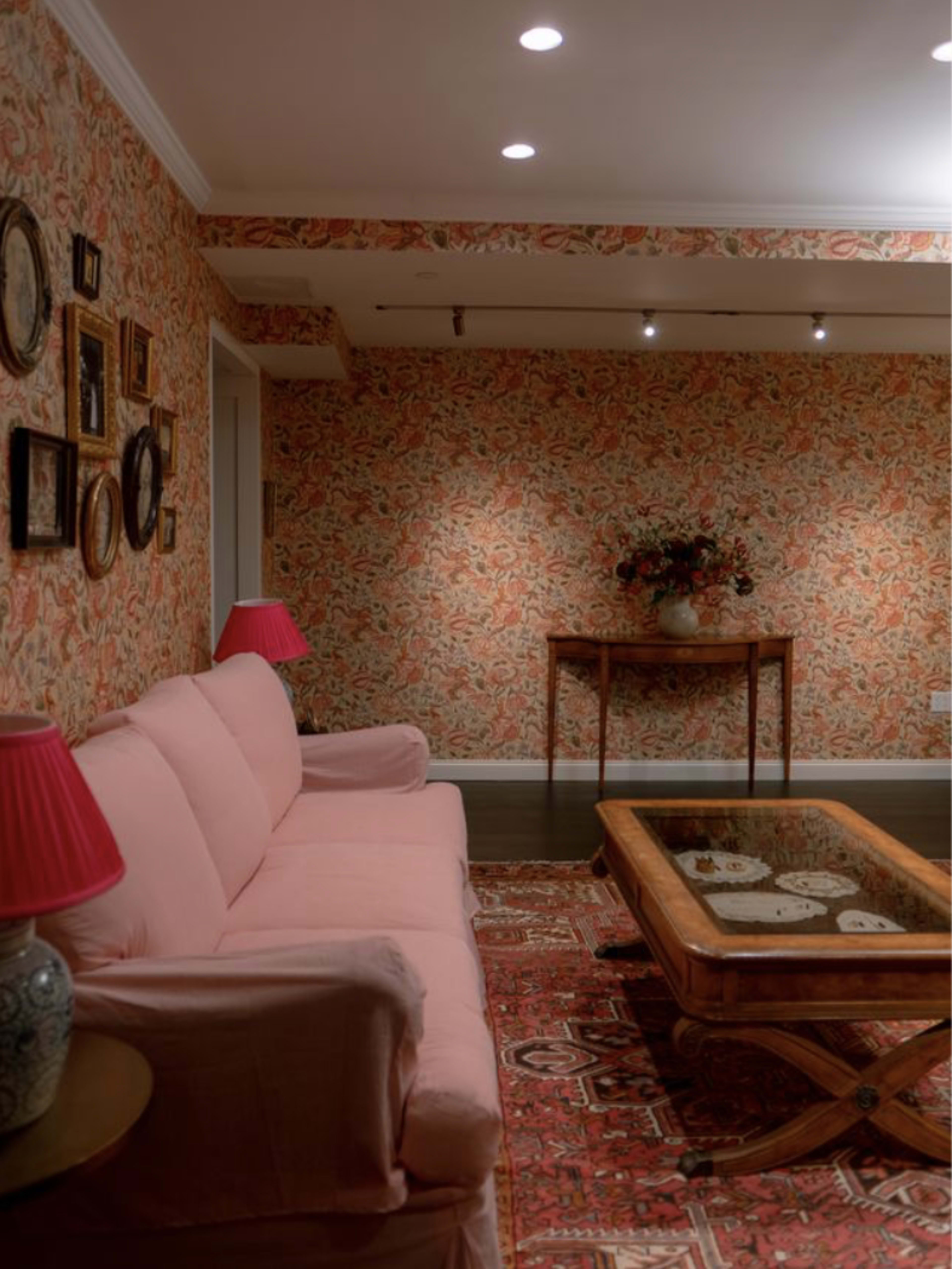The image shows a living room with a pink sofa, a wooden coffee table, and floral wallpaper adorned with framed pictures and a vase of flowers.