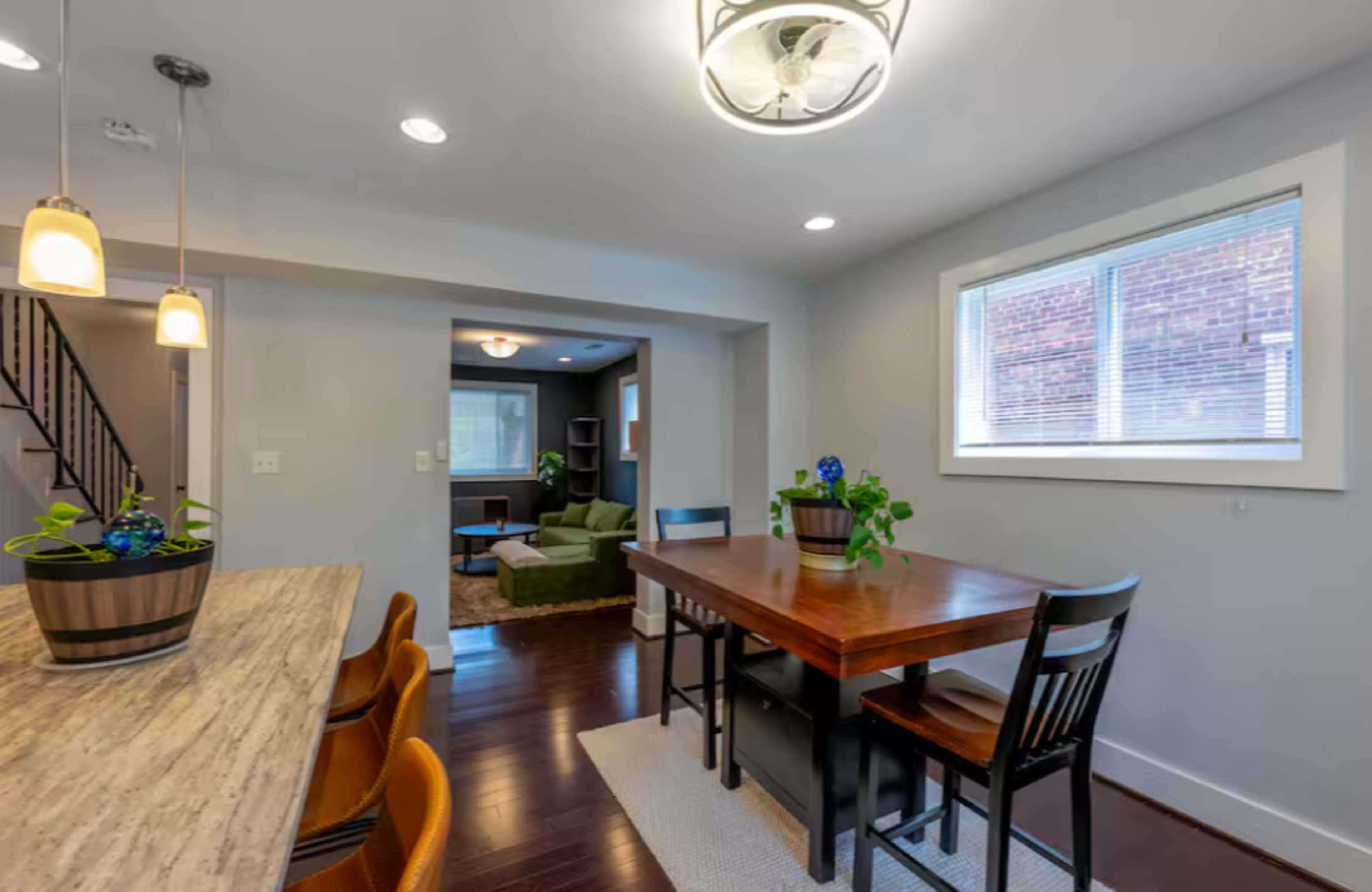 The image shows a dining area with a wooden table and chairs, illuminated by recessed lighting, connected to a living room visible in the background.
