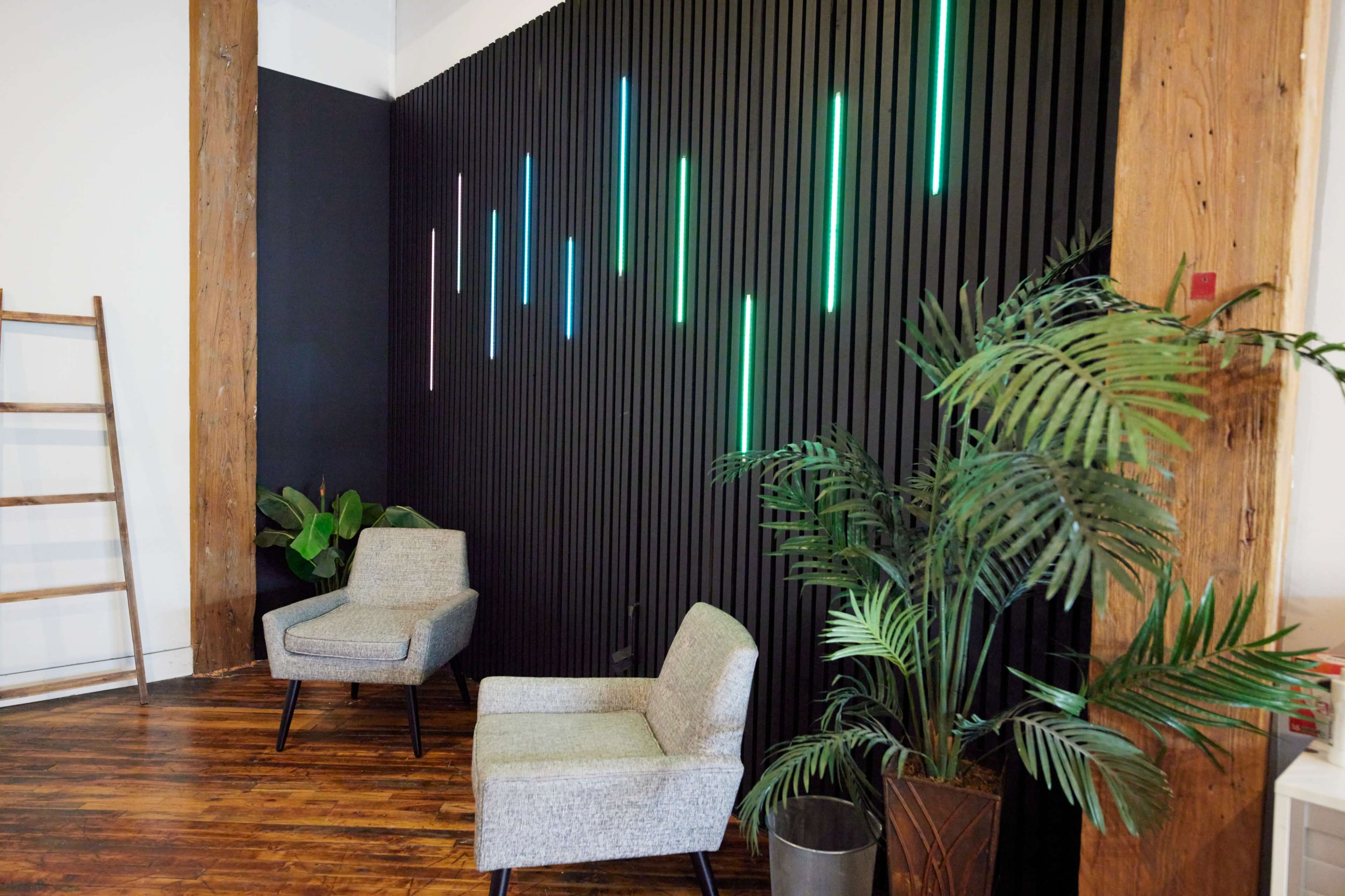The image shows two grey armchairs positioned next to a potted plant in front of a textured black wall with vertical, illuminated light strips in varying colors.