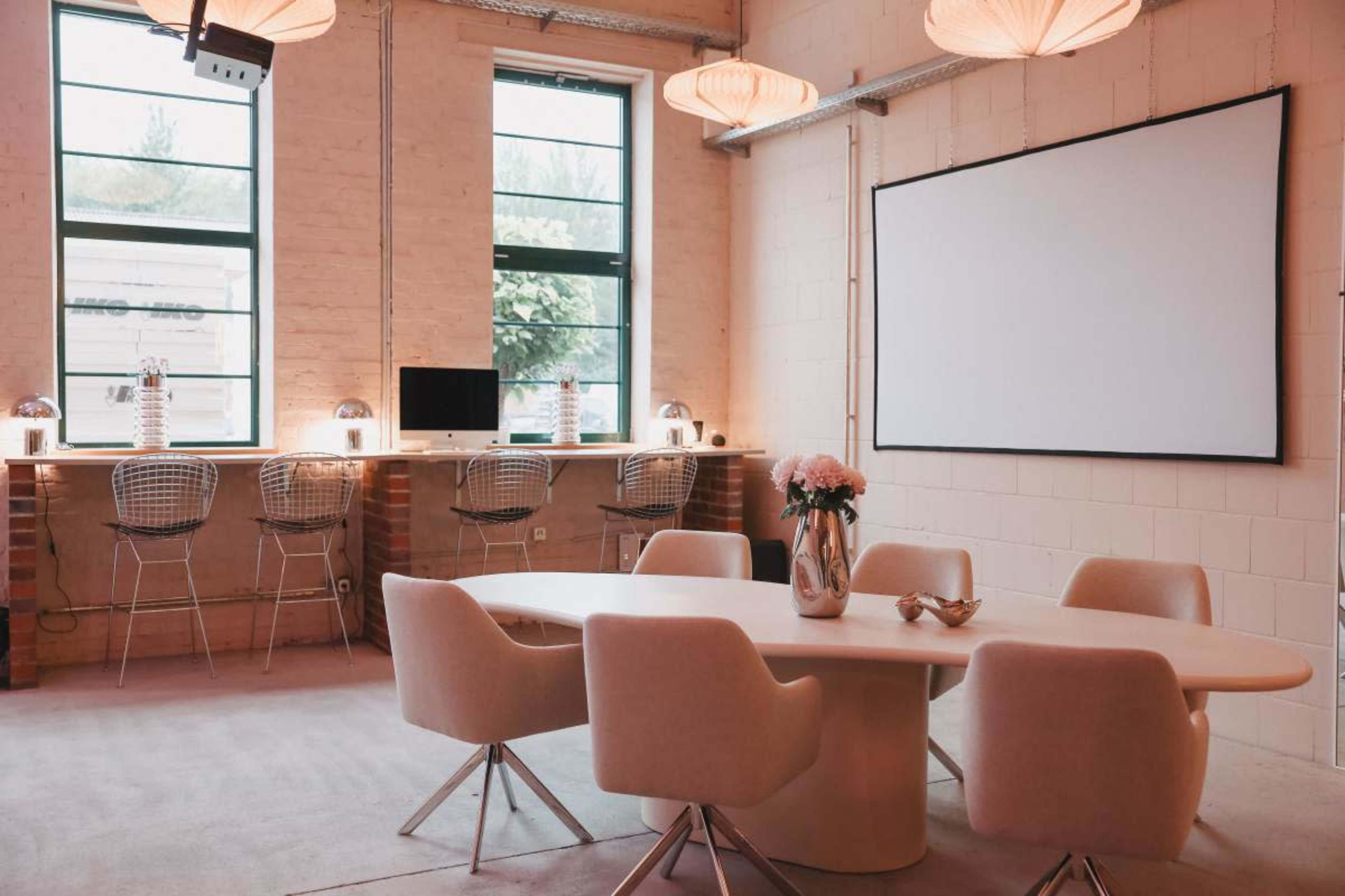 A modern meeting room features a round table surrounded by white chairs, with a large screen on one wall and bar-style seating near windows.