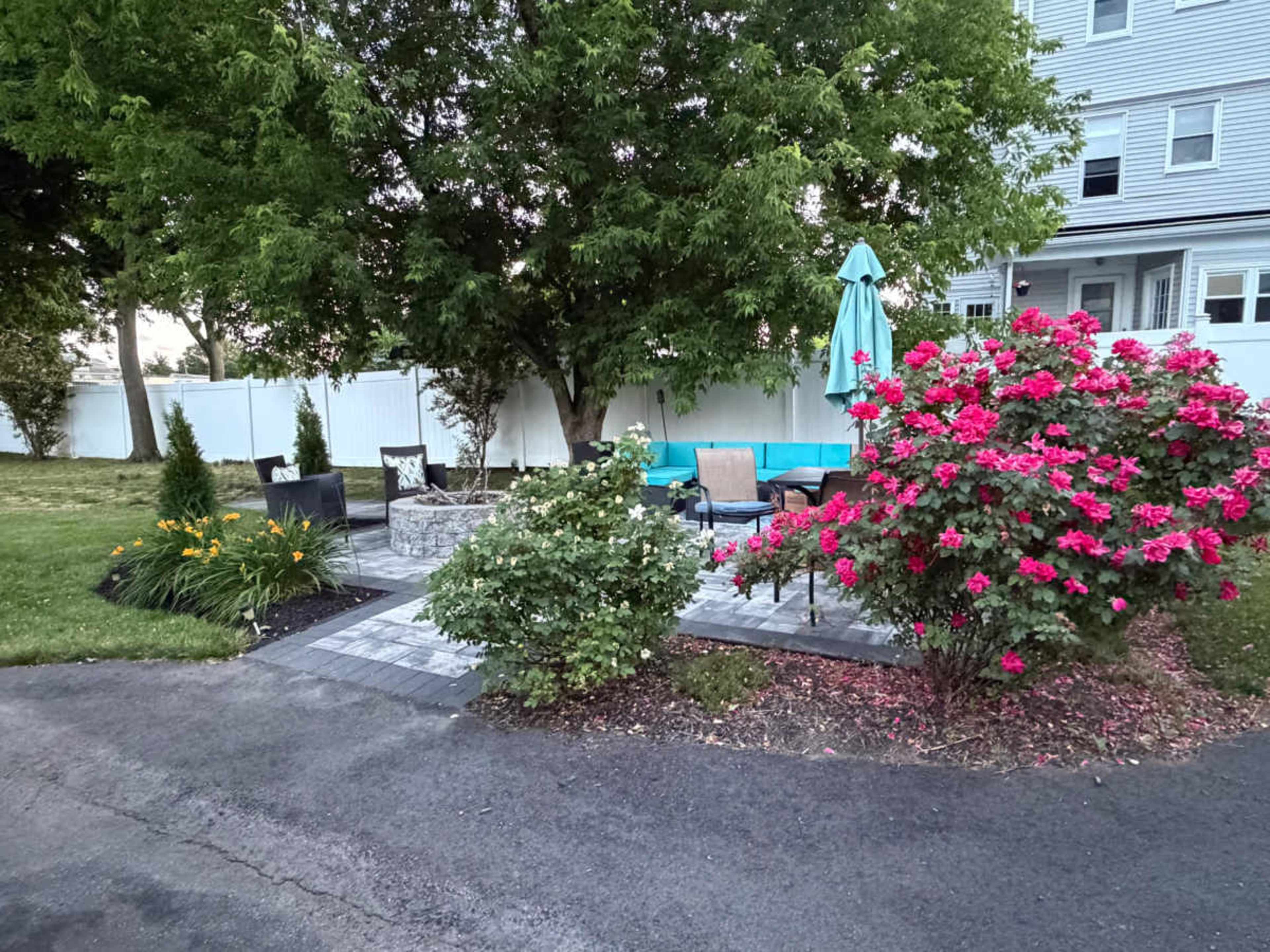 The image shows a landscaped outdoor seating area featuring a stone patio, surrounded by colorful flowers, shrubs, and an umbrella, with a grassy area nearby.