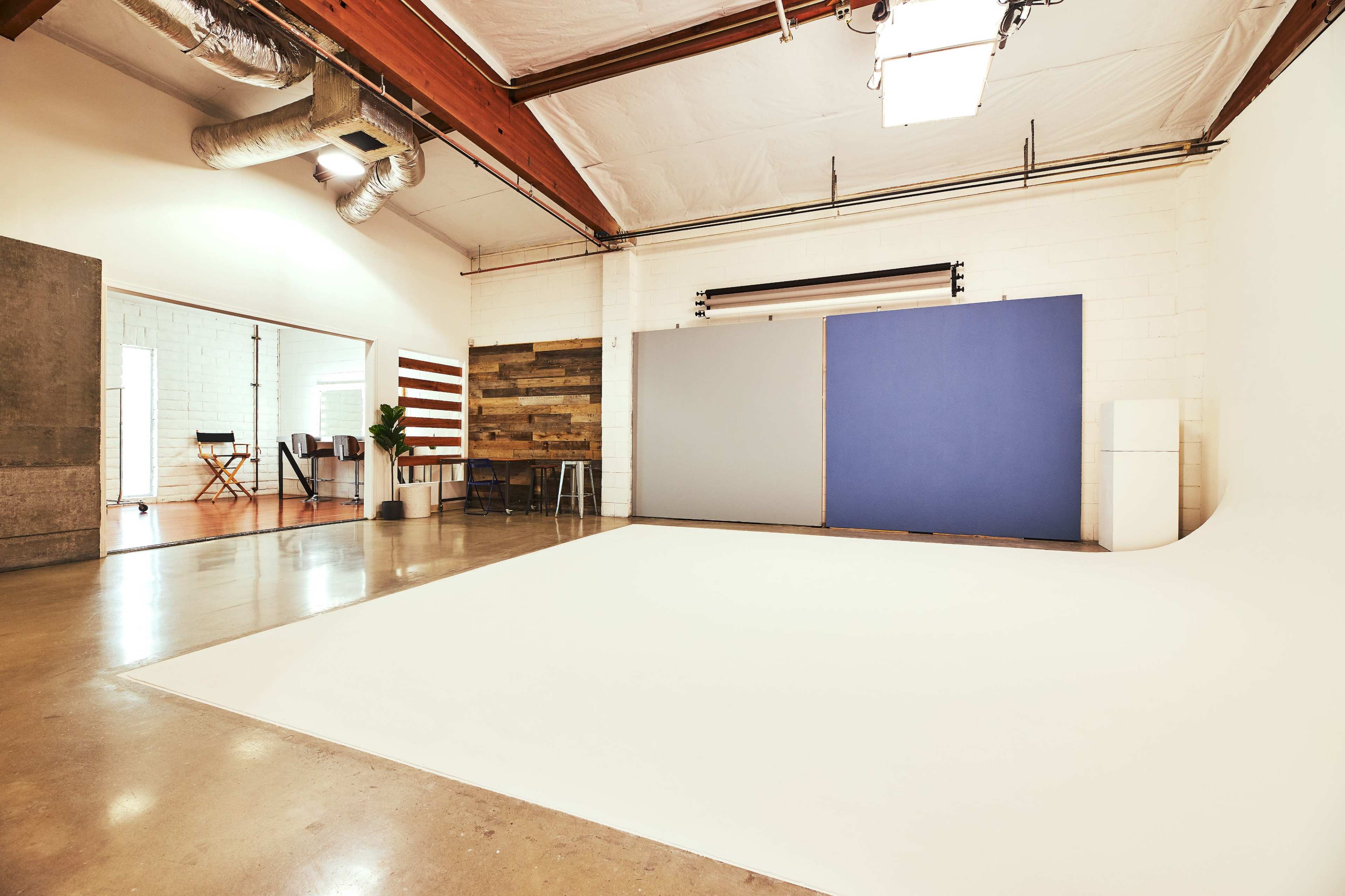 A spacious photography studio with a white backdrop, wooden paneling, and a refrigerator, featuring industrial elements such as exposed ductwork and wooden beams.