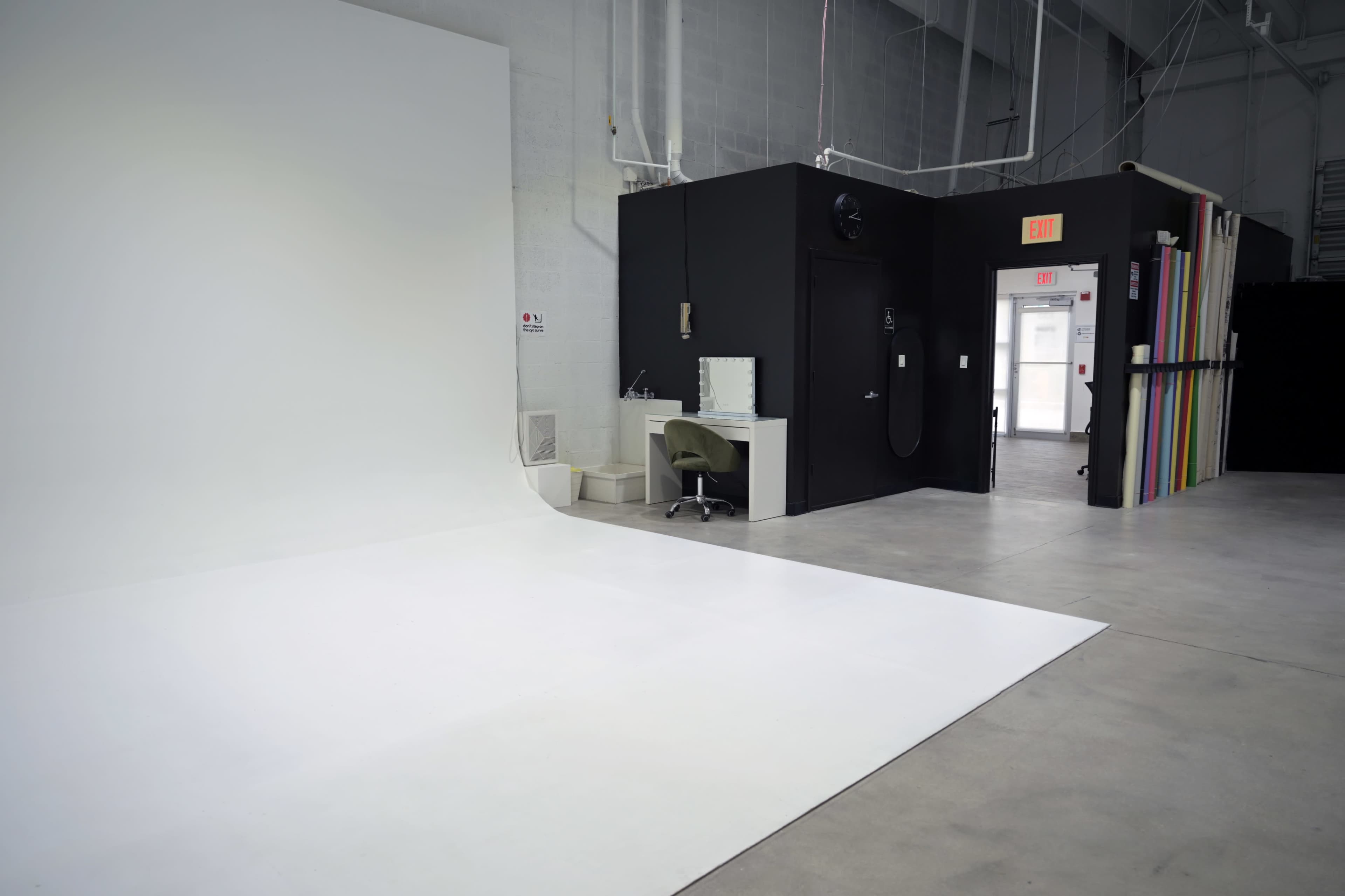 The image shows a spacious photography studio featuring a large white backdrop on one side and a small black entrance with a mirror and chair on the other.