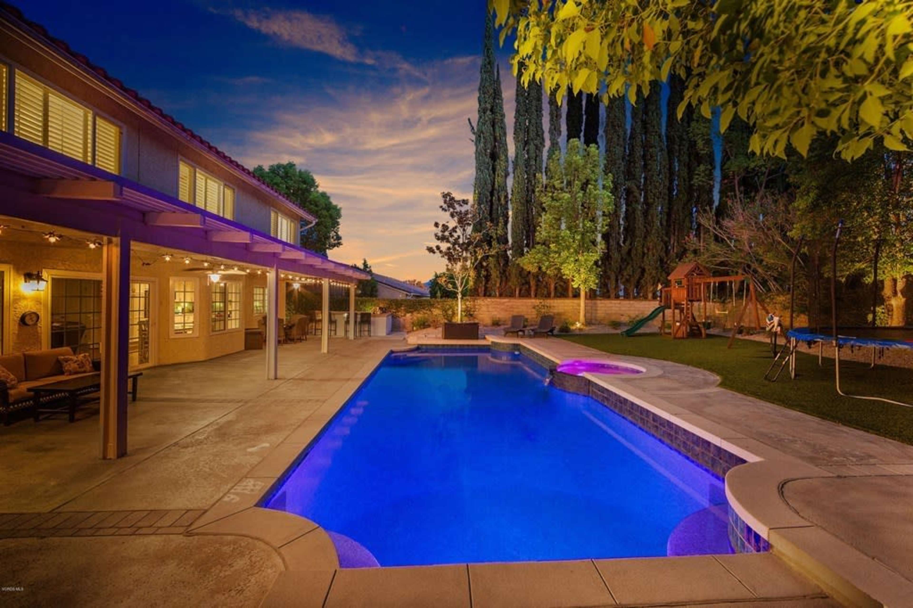 The image shows a well-lit backyard featuring a swimming pool with blue lighting, surrounded by trees and a playset.