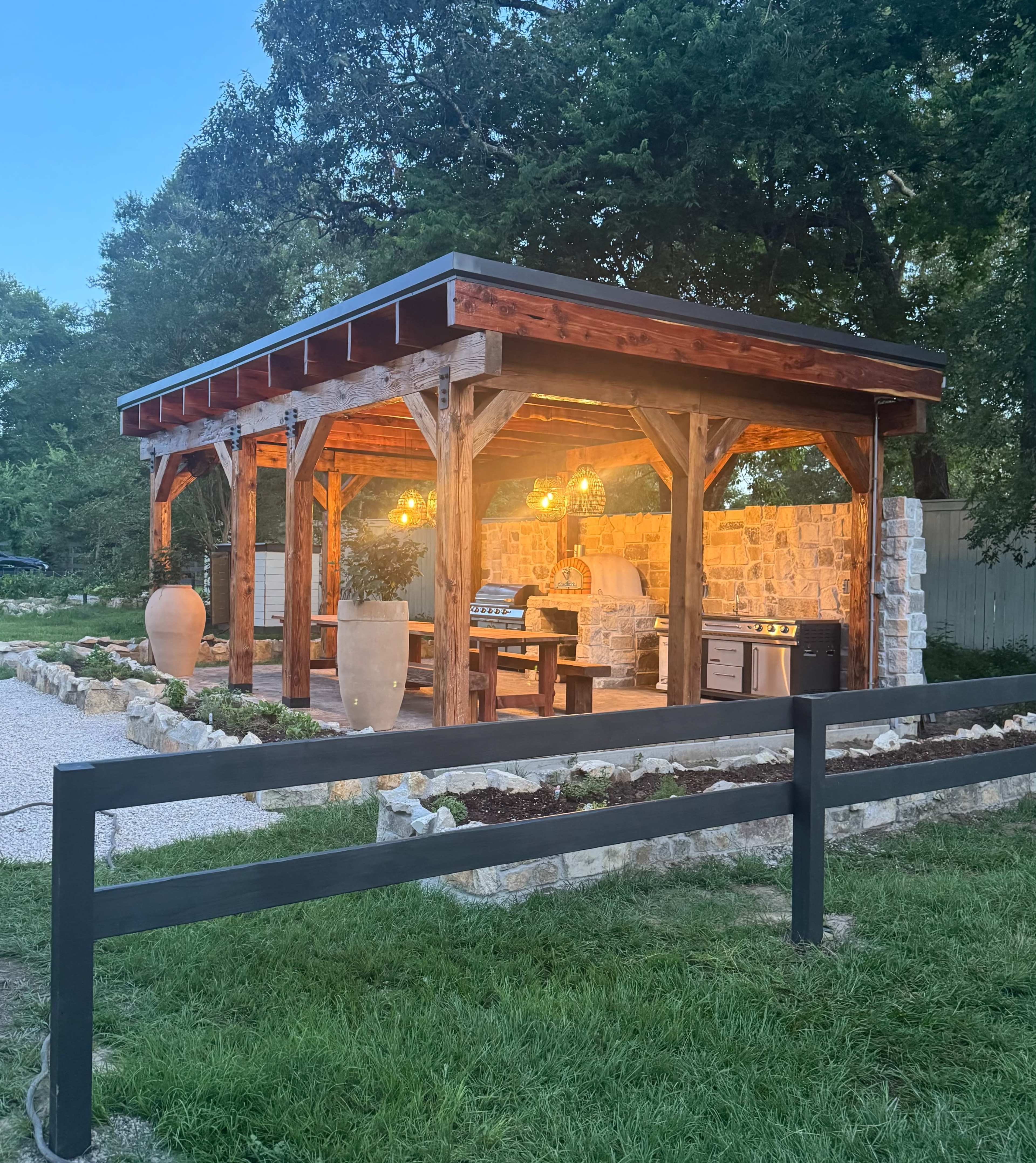 The image shows a wooden outdoor pavilion with stone walls, equipped with a barbecue grill and seating, illuminated by hanging lights.
