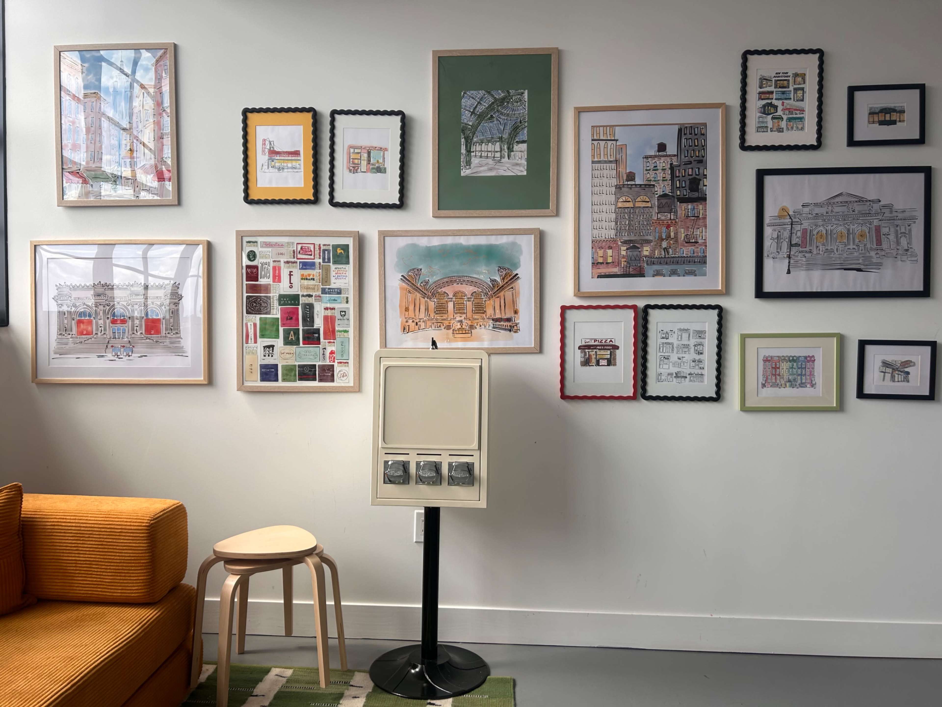 The image shows a wall decorated with various framed artworks, including illustrations and prints, surrounded by a cozy seating area with an orange couch and a wooden stool.