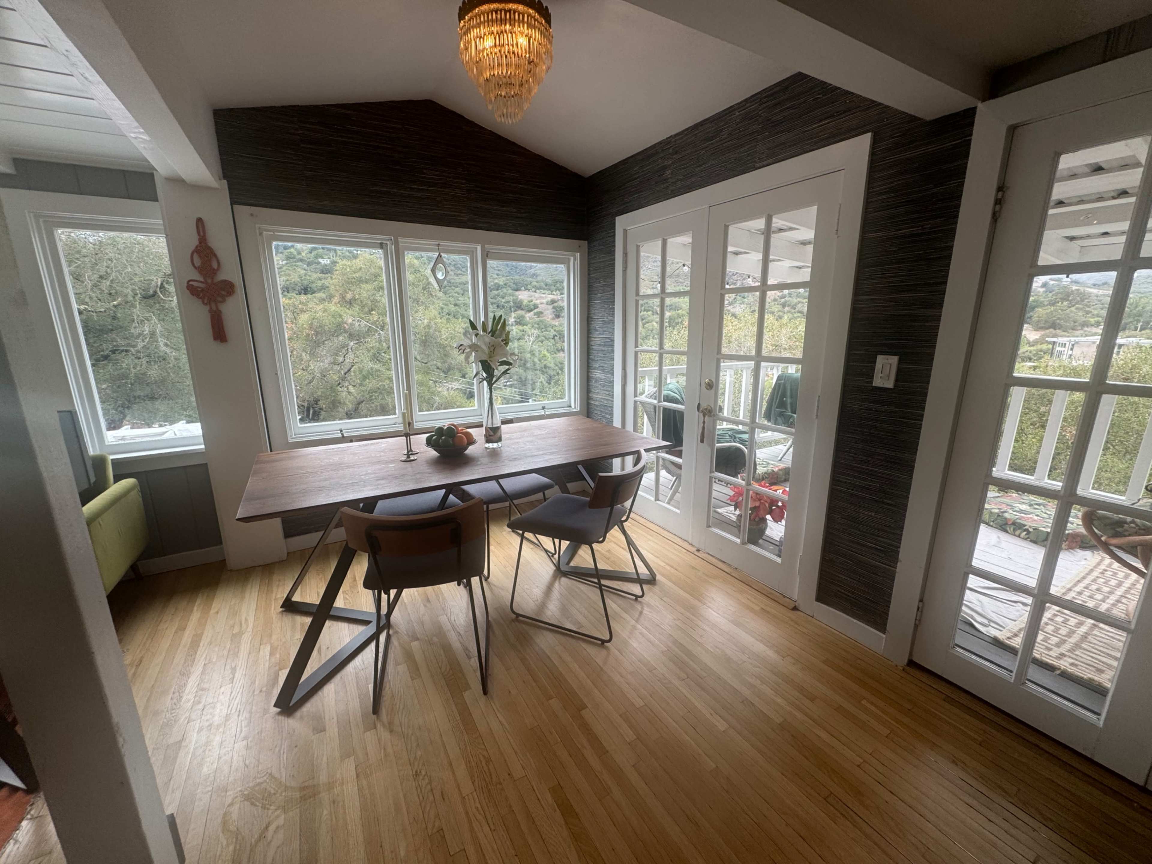 The image shows a dining area with a wooden table surrounded by chairs, large windows offering a view of greenery, and a door leading to a patio.