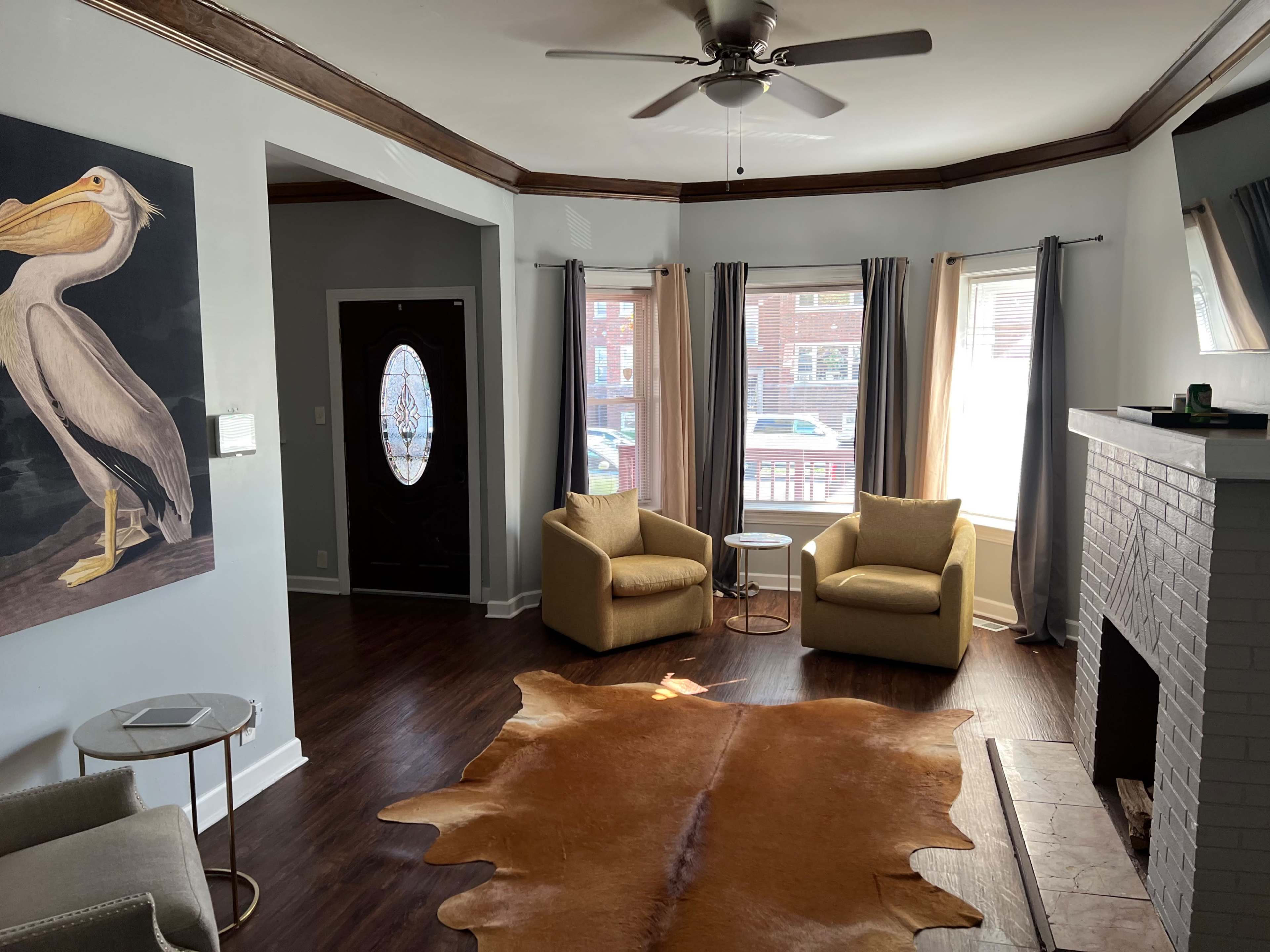 The living room features two yellow armchairs, a round side table, and a tan cowhide rug, with large windows allowing natural light and a pelican painting on the wall.