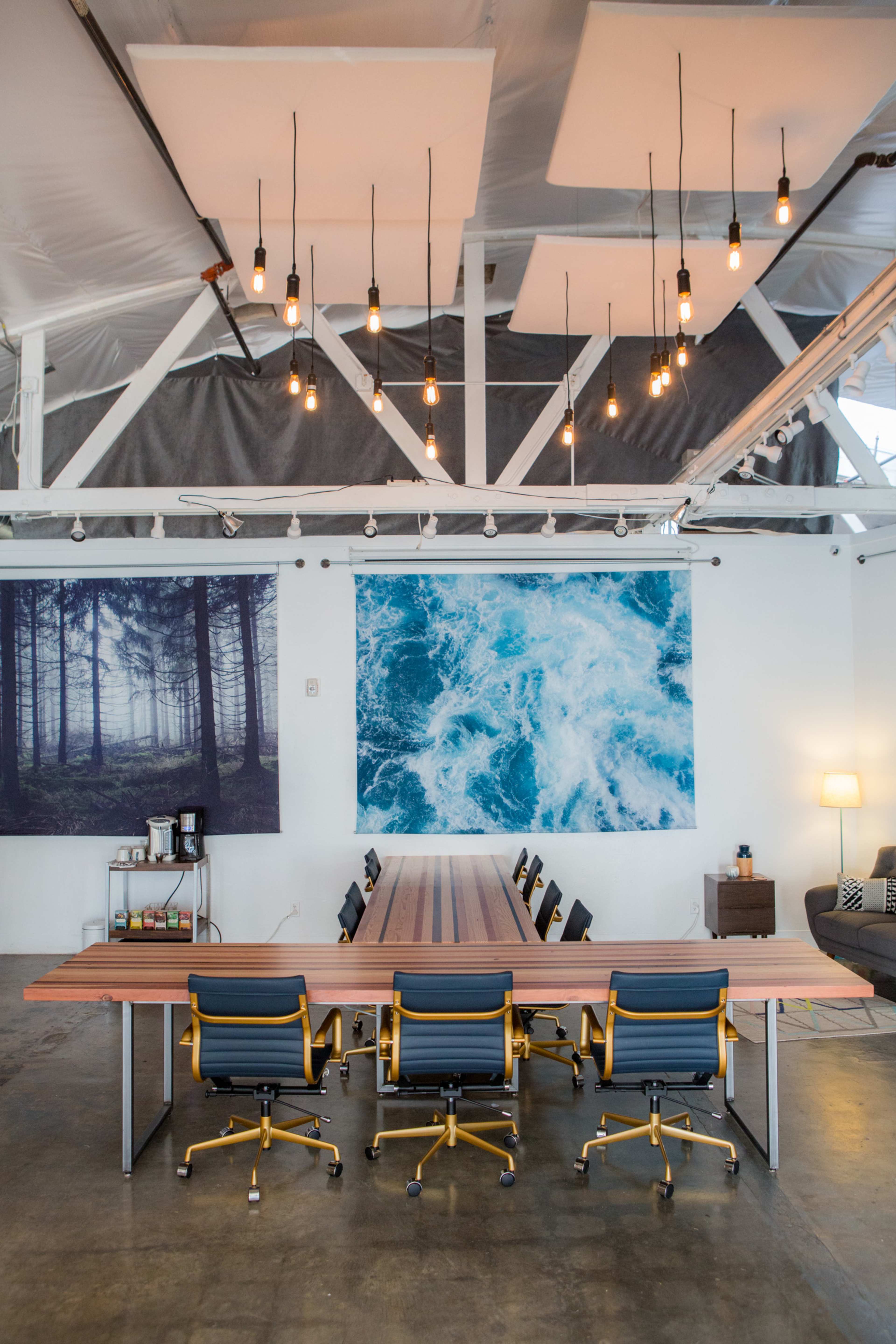 A long wooden conference table with six yellow chairs is set up in a spacious, well-lit room featuring large art prints of a forest and ocean on the walls.