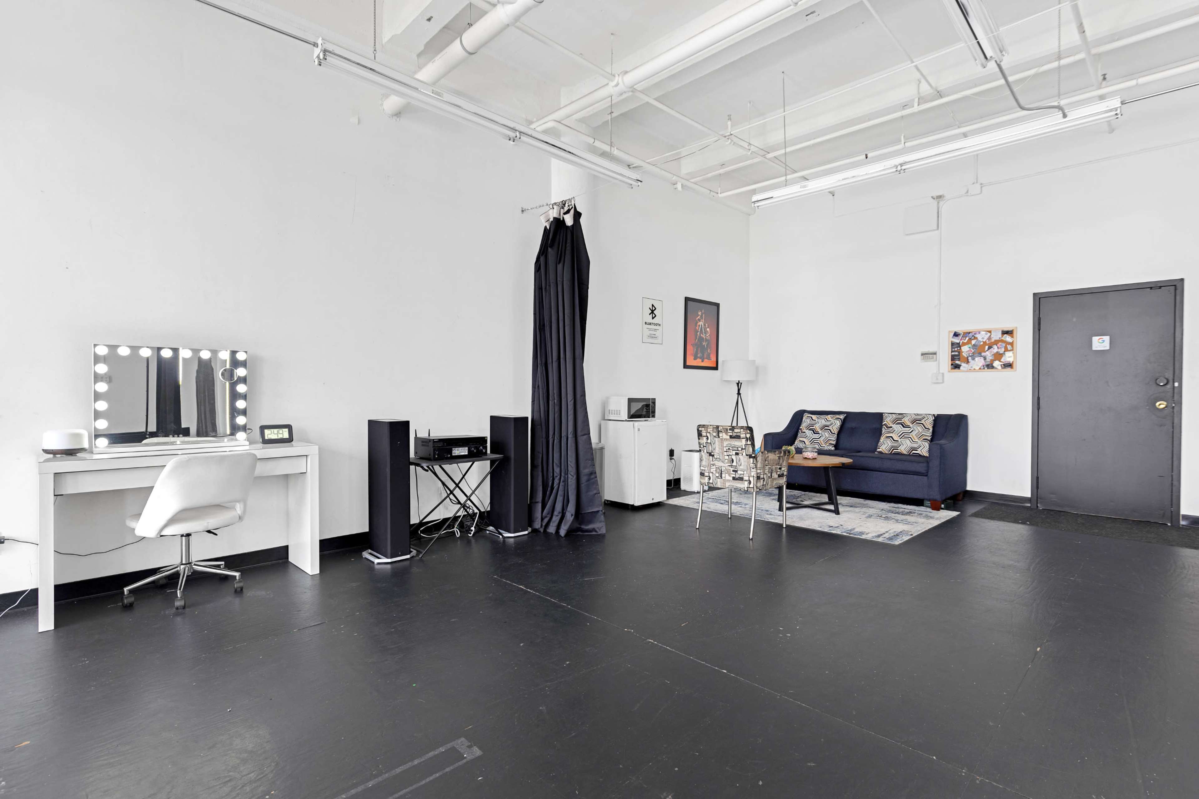 The image shows a minimalistic studio space featuring a white desk with a mirror, a black curtain, a blue sofa with patterned cushions, and a black door.