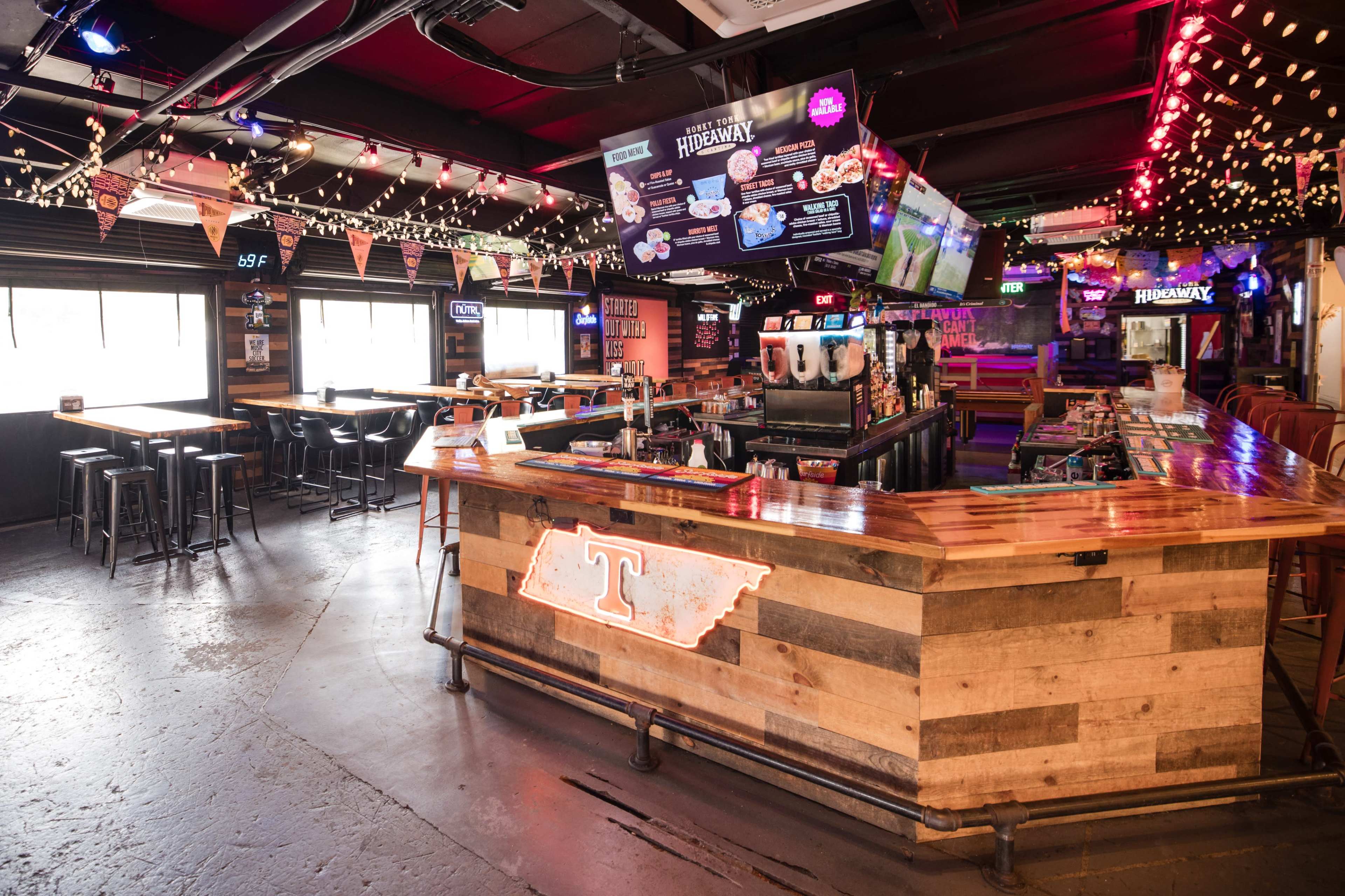 The image shows a bar area with wooden countertops, colorful lighting, and several televisions displaying information and sports.