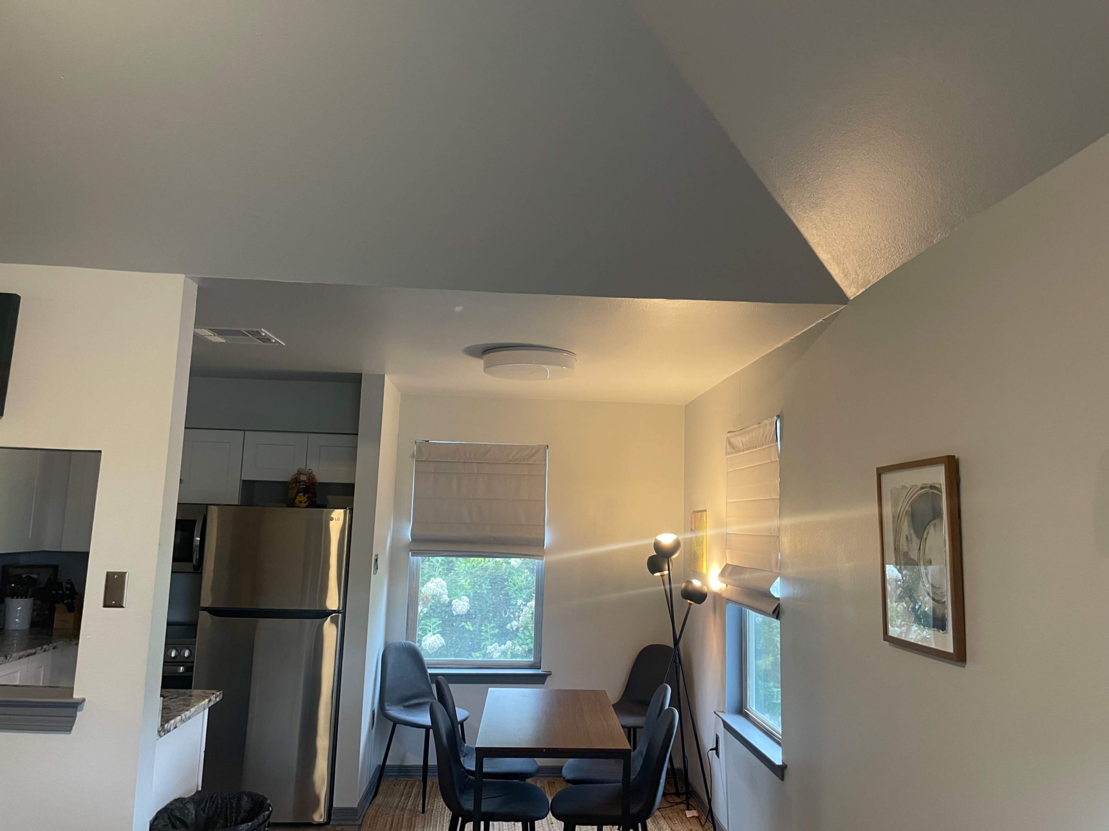 The image shows a bright, modern dining area with a table, four chairs, and a large window beside a stainless steel refrigerator.