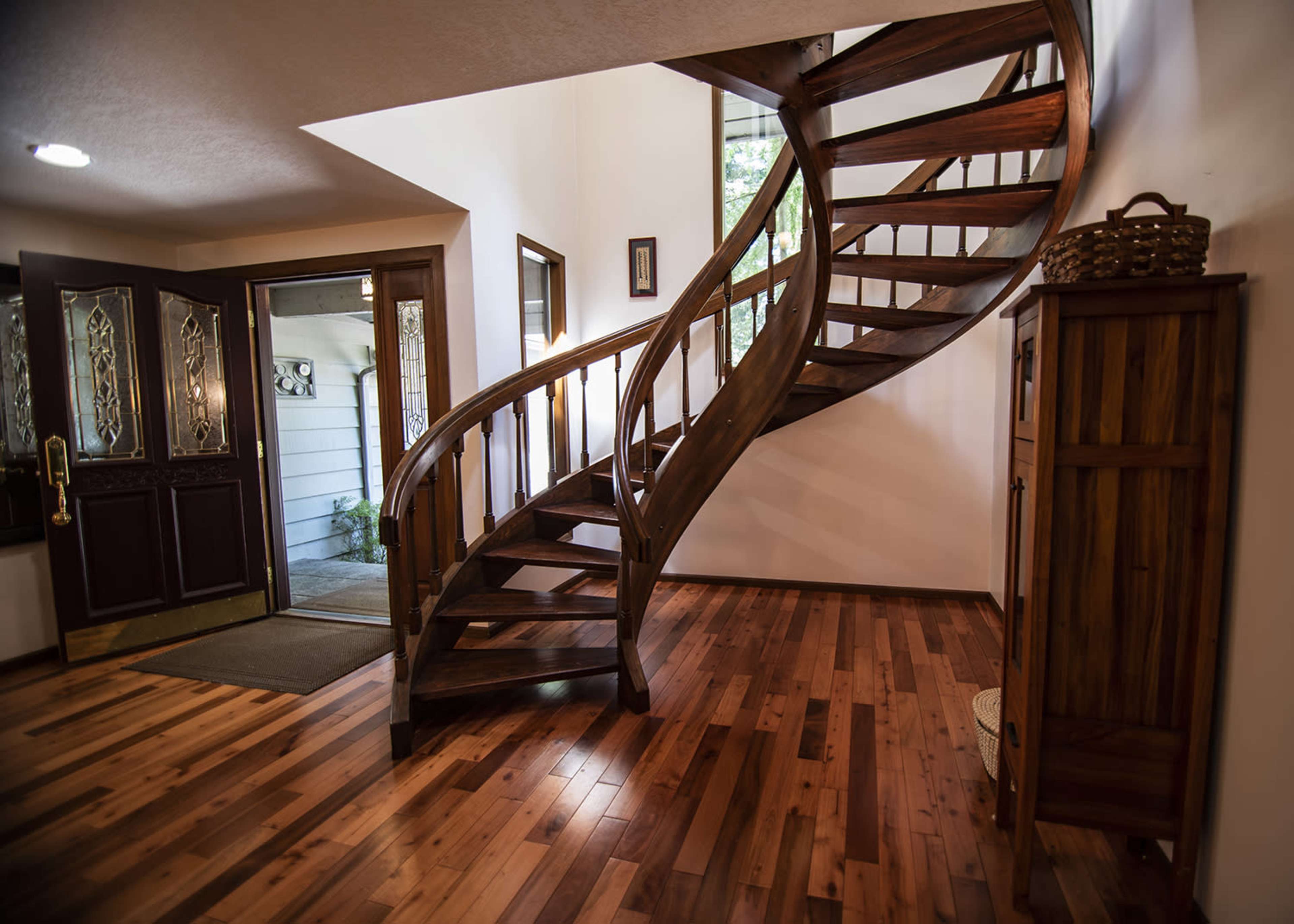 A wooden spiral staircase leading to an upper level, set within a warmly lit entryway that includes a front door and a wooden cabinet.