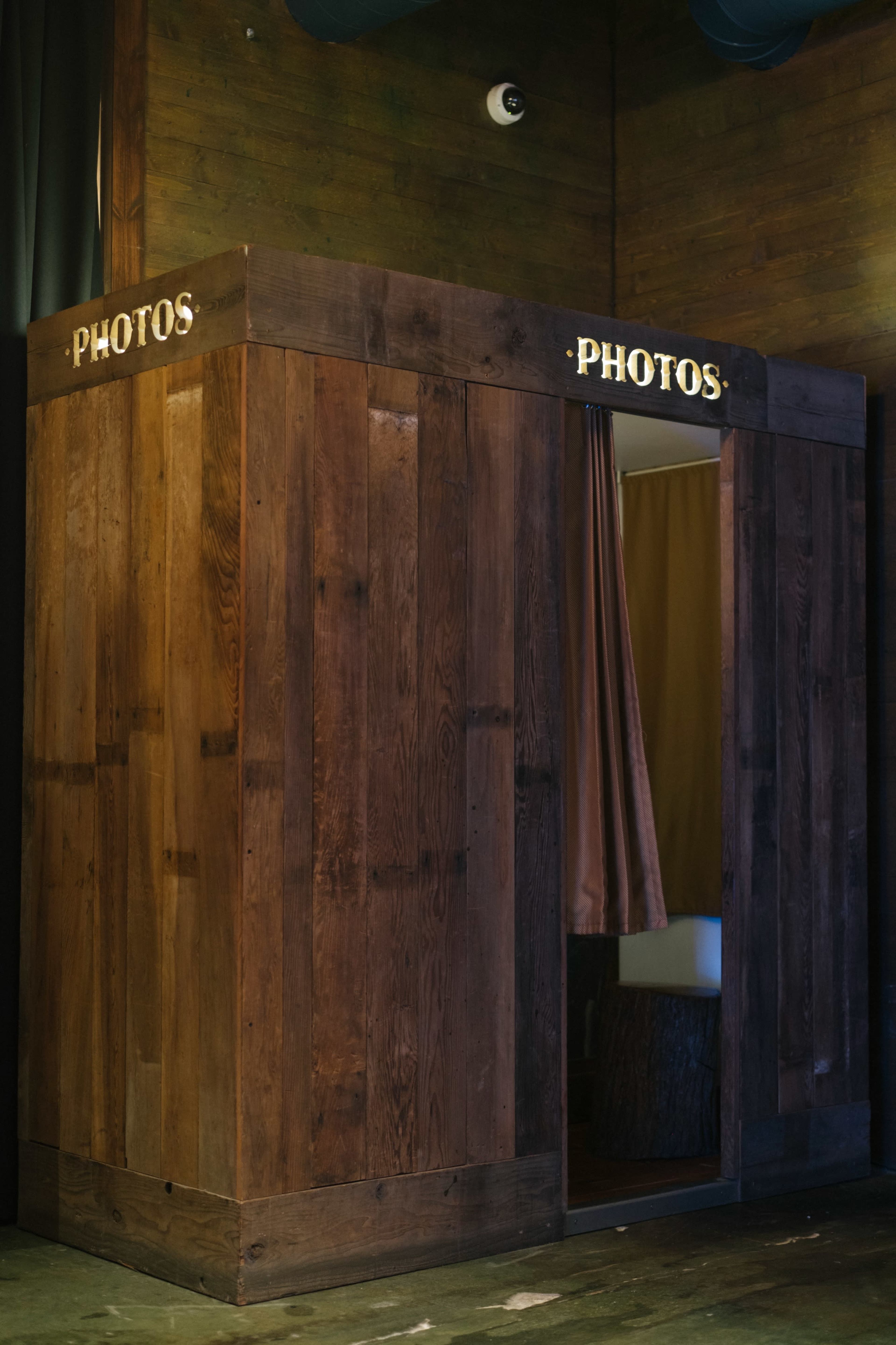 The image shows a wooden photo booth with "PHOTOS" written on the top, featuring an open entrance and a curtain inside.
