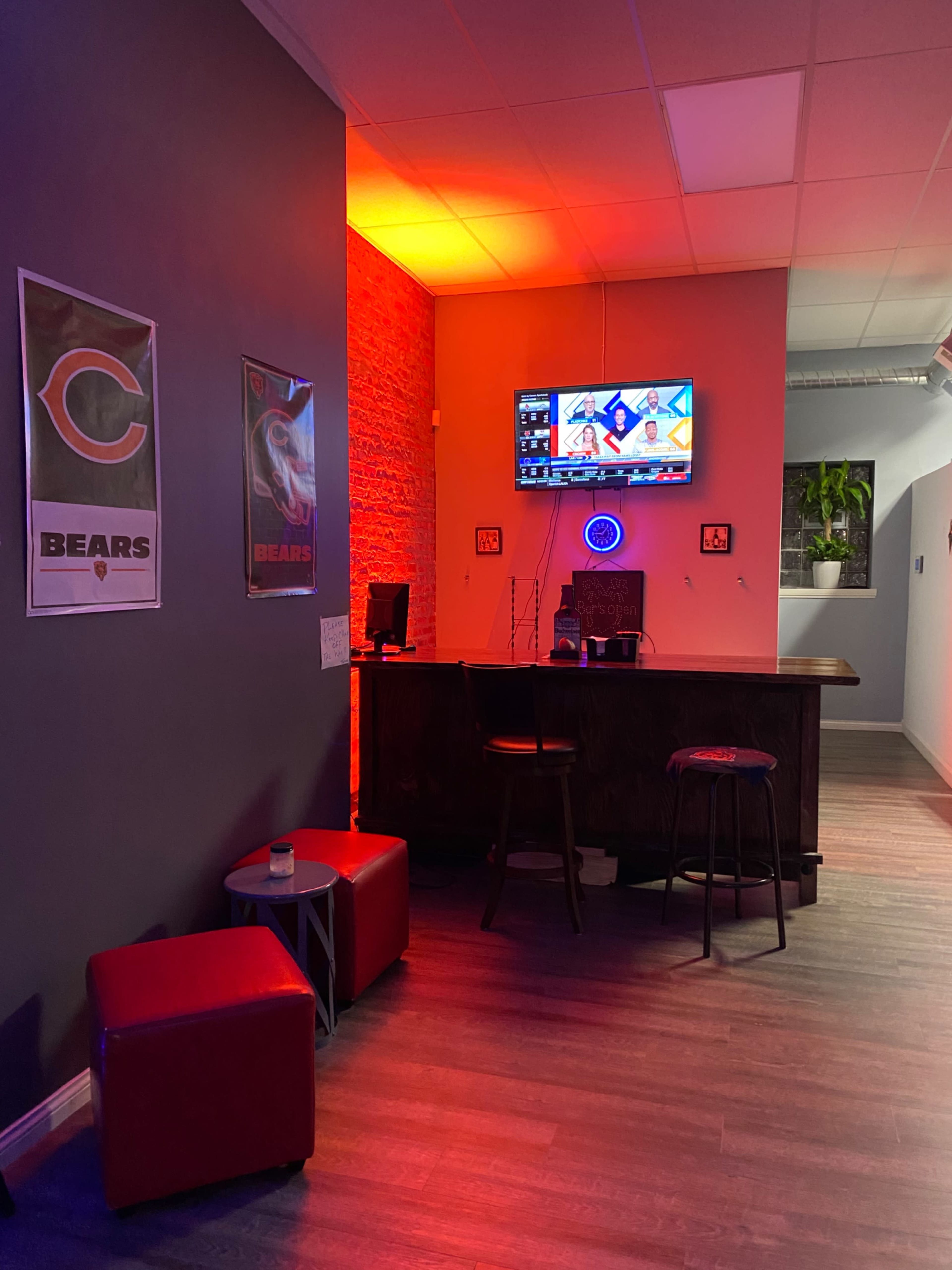The image shows a small bar area with red stools, a countertop, and a television displaying a menu, illuminated by orange and red lighting.