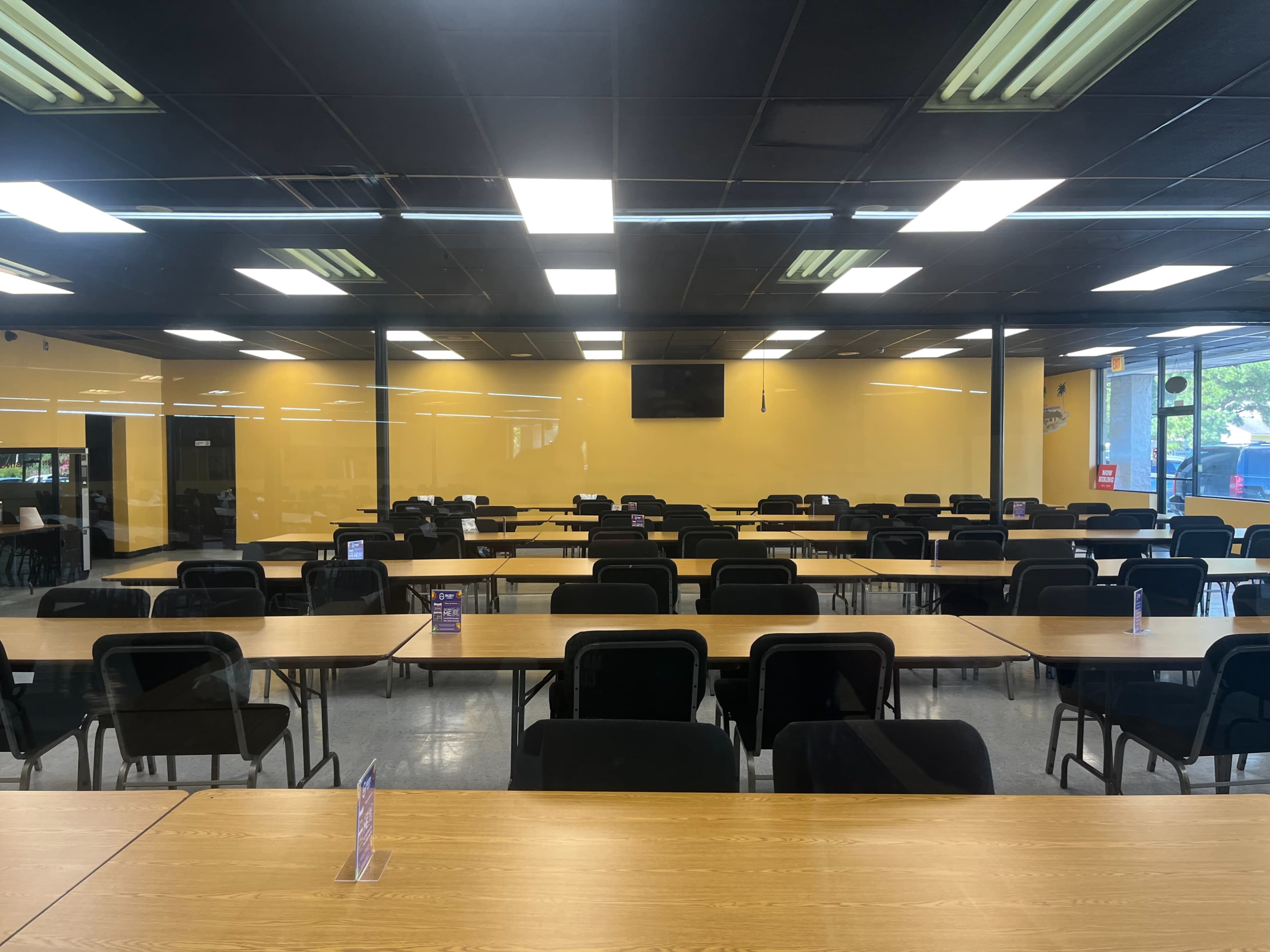 A spacious dining area features rows of wooden tables with black chairs, illuminated by fluorescent lights.