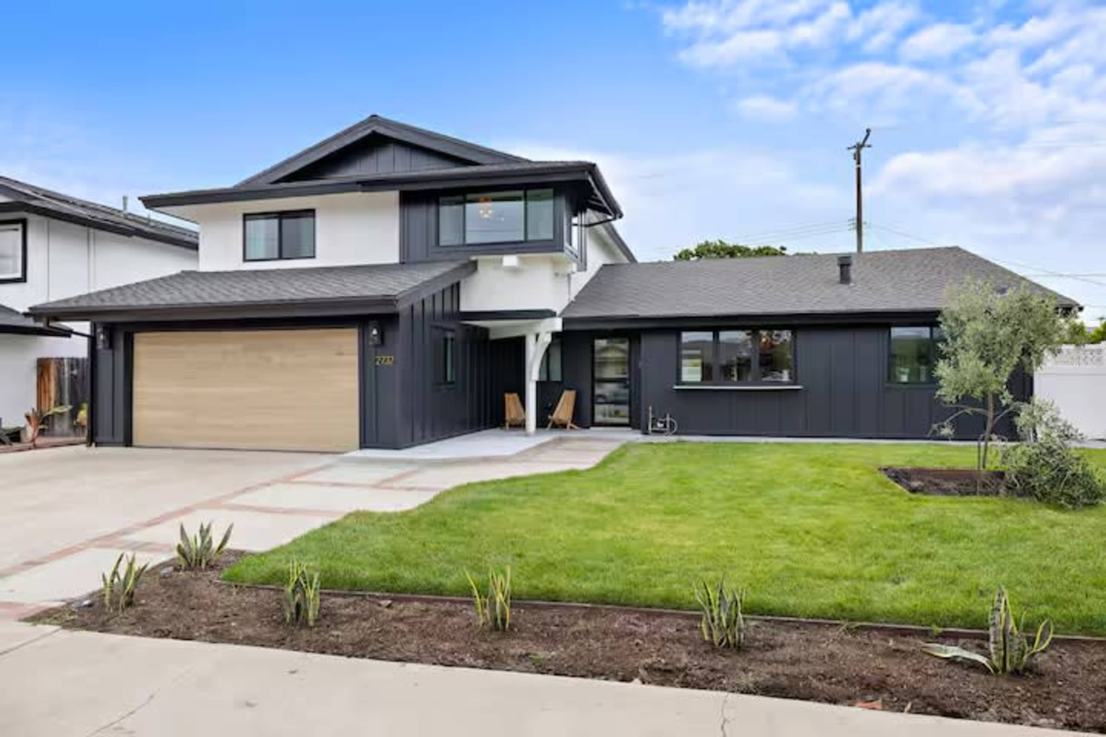 A modern two-story house features a combination of dark siding and a light wooden garage door, set on a manicured lawn with landscaping.