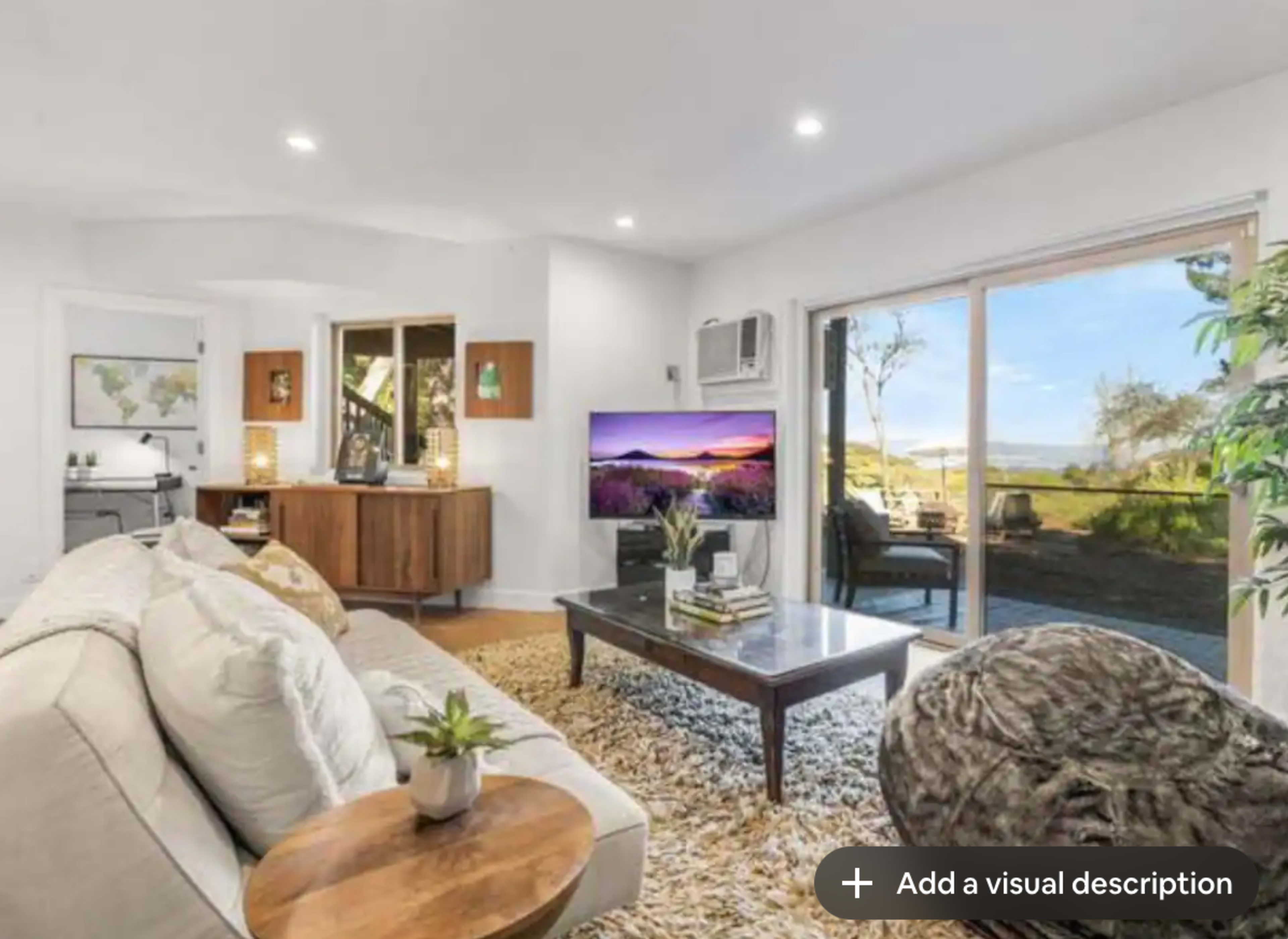 A modern living room with a beige sofa, a wooden coffee table, and a television displaying a landscape, with large windows providing a view of the outdoors.