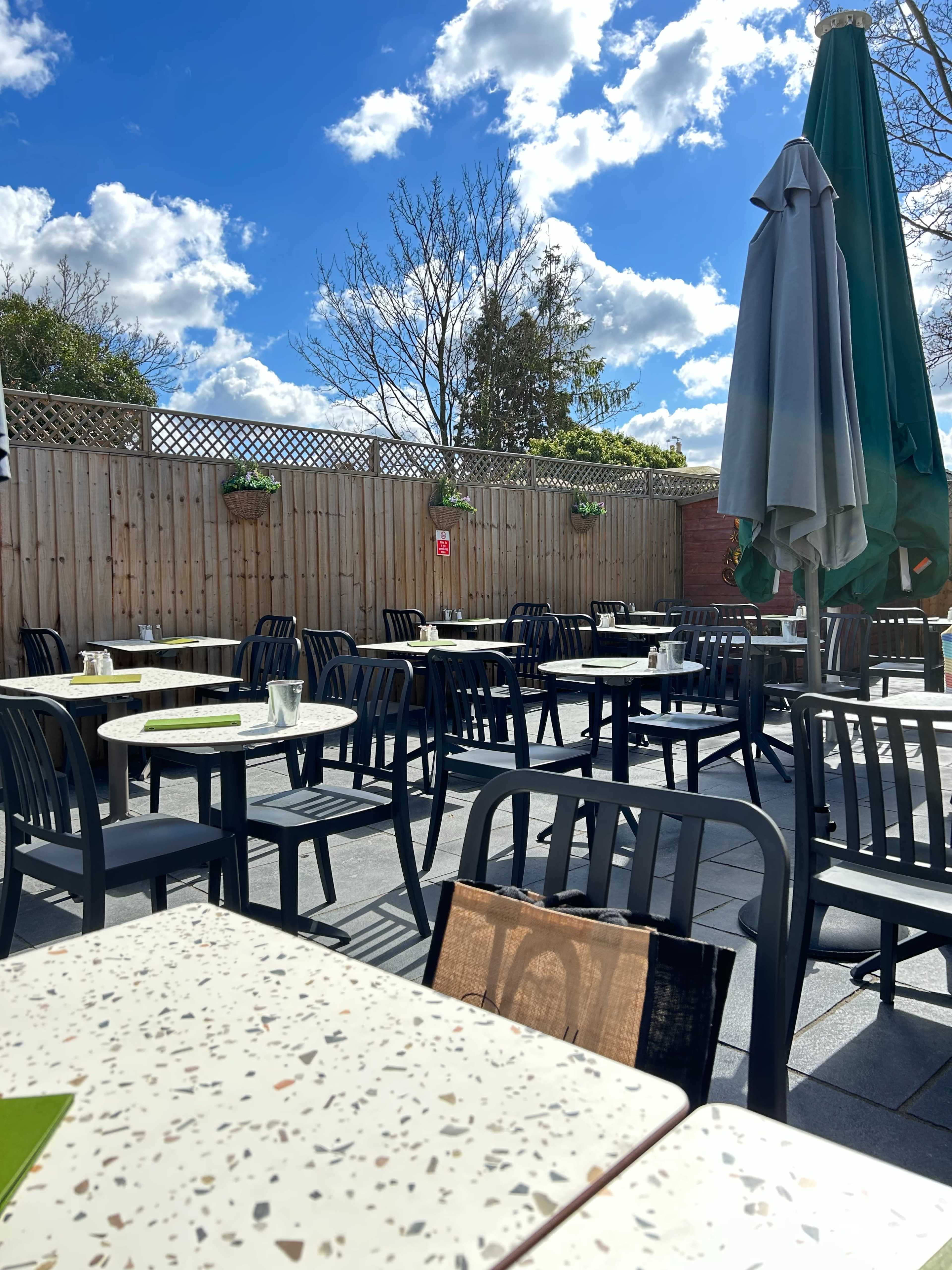 A patio area at a restaurant features several empty tables and chairs under a bright sky with scattered clouds.