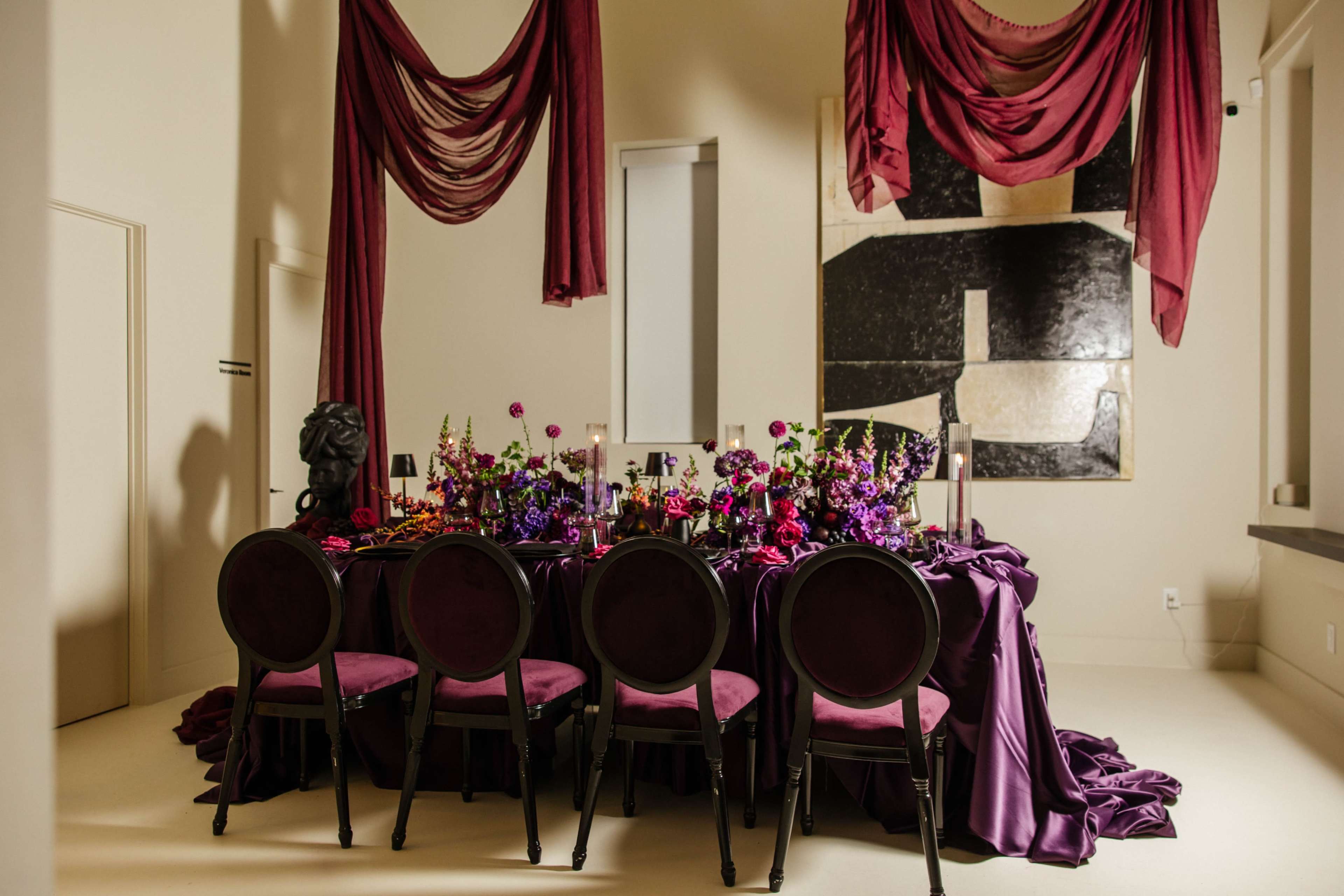 A long table set for a formal dinner is adorned with deep purple flowers and draped fabrics, surrounded by elegant black chairs.