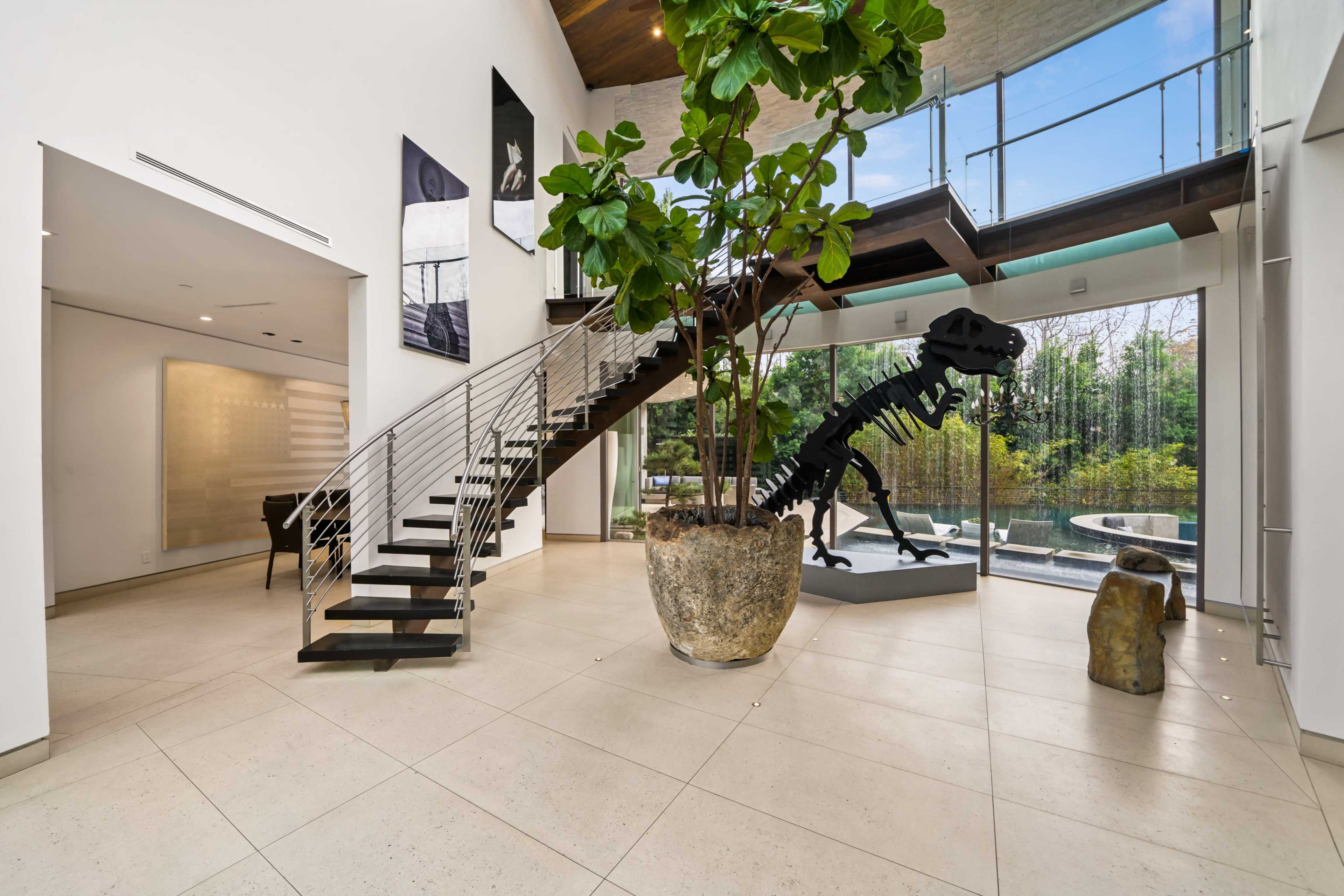 A modern entrance hall featuring a staircase, a large potted plant, and a dinosaur skeleton sculpture, with large windows overlooking a garden.