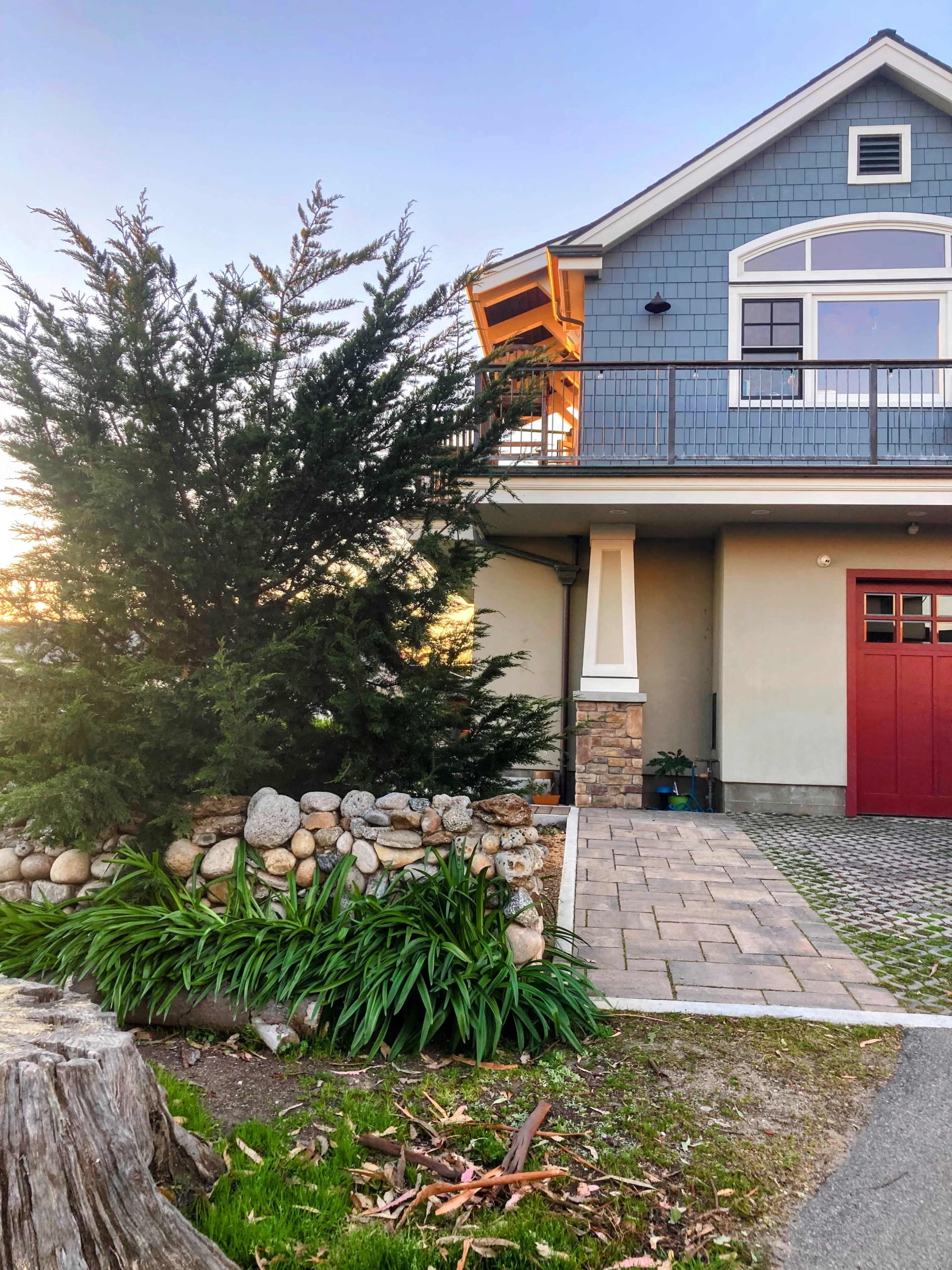 A house with a blue exterior, stone accents, and a red door is surrounded by landscaped greenery and a stone wall.