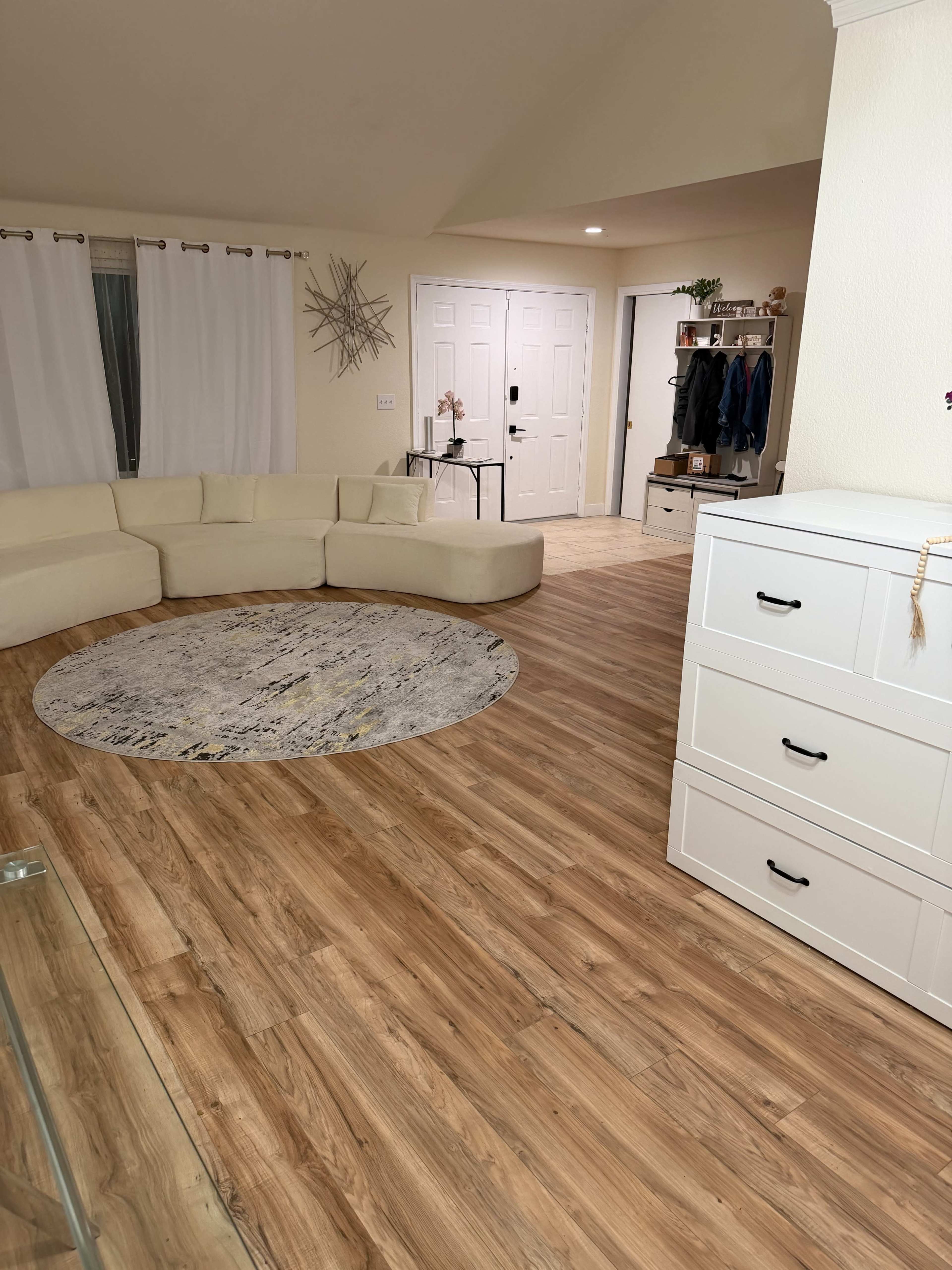 The image shows a neatly arranged living room with a curved beige sofa, a round area rug, light wood flooring, and a white dresser against the wall.